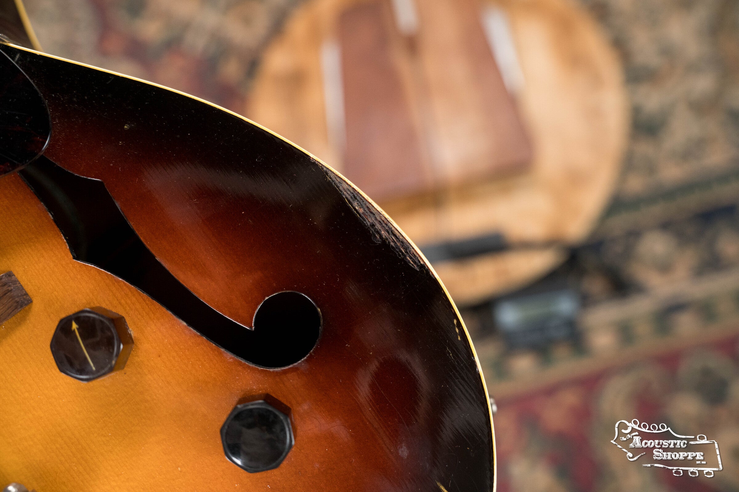 Close-up of a sunburst-finish (Used) 1942 Gibson ES-125 hollow-body guitar by Gibson, highlighting tuning pegs and an f-hole, with a blurred instrument and patterned rug in the background. Acoustic Shoppe logo appears in the lower right corner.