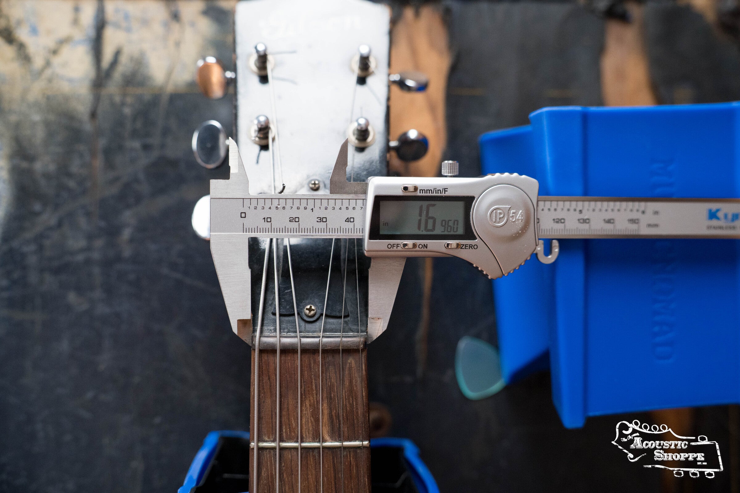 A digital caliper measures the nut width of a (Used) 1942 Gibson ES-125 by Gibson, displaying 1.695 inches. The guitar rests on blue supports atop a workbench, with The Acoustic Shoppe logo in the lower right corner.