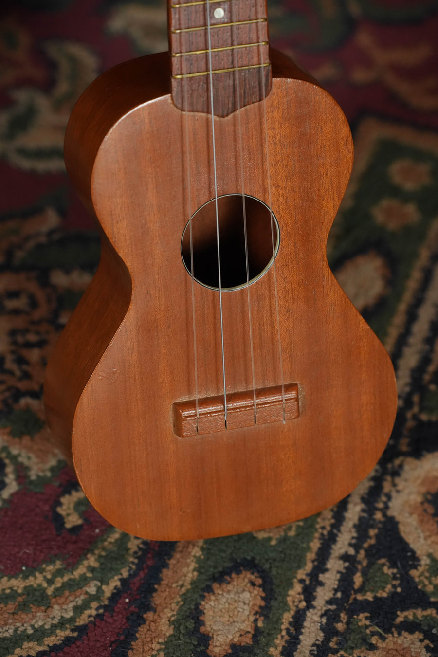 A close-up of a (Used) 1960s Vintage Airline Mahogany Soprano Ukulele from The Acoustic Shoppe, featuring four strings and set against a patterned, multicolored carpet background.