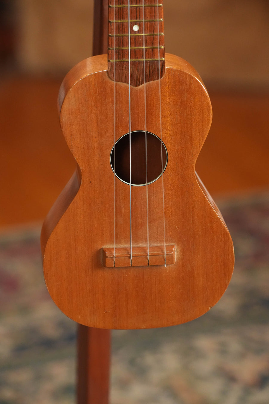 A close-up of a (Used) 1960s Vintage Airline Mahogany Soprano Ukulele from The Acoustic Shoppe, shown upright with four strings. The softly blurred background features a patterned rug and warm-toned floor.