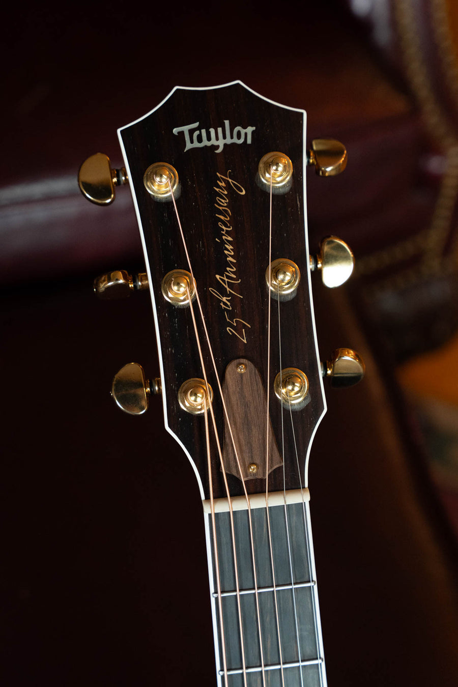 Close-up of a (Used) 1999 Taylor 814-BCE 25th Anniversary Sitka/Rosewood Grand Auditorium Guitar headstock with gold tuning pegs, script "25th Anniversary" below the Taylor Guitars logo, blurred brown leather background.