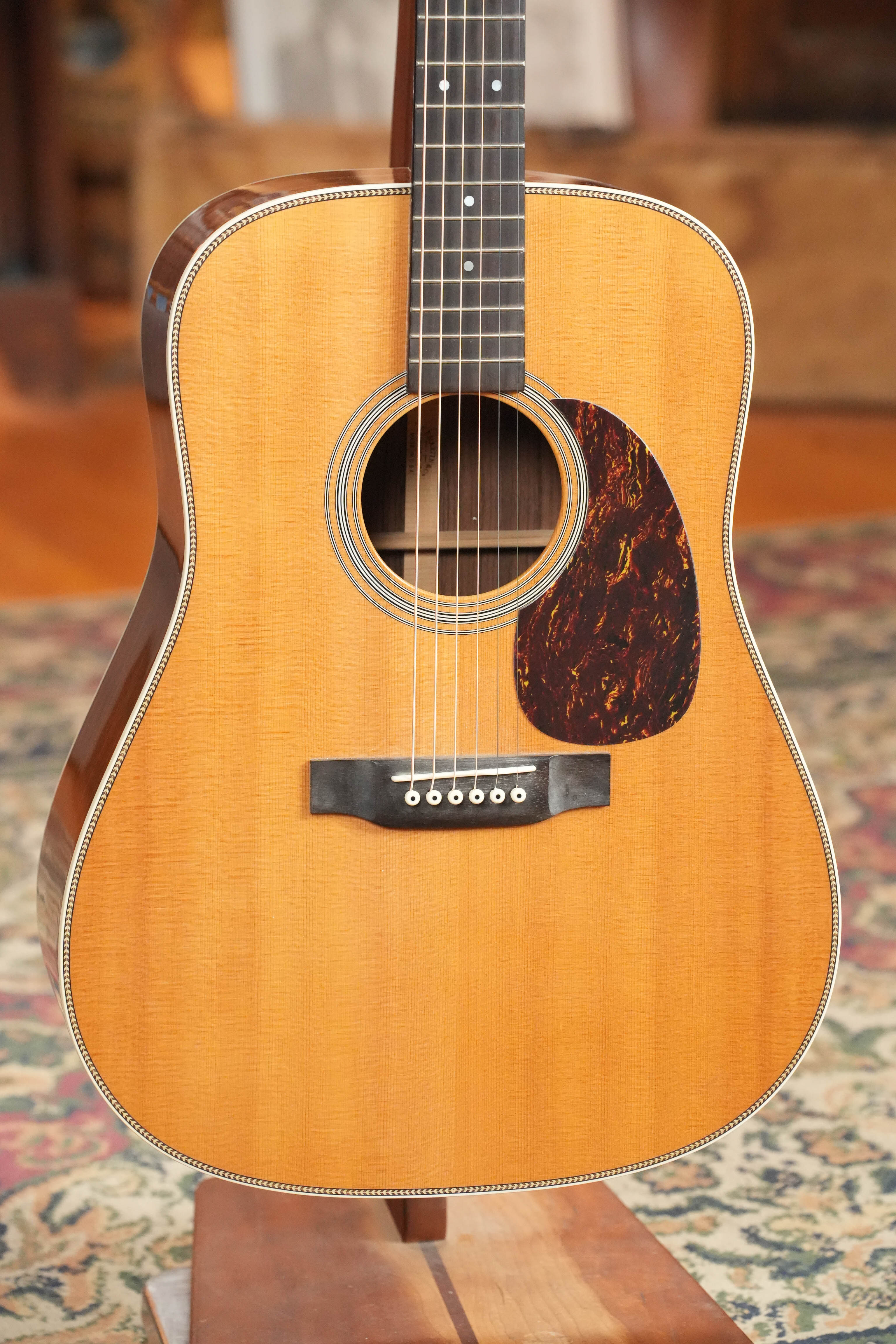 A close-up of a (Used) 2007 Martin & Co. HD-28 dreadnought acoustic guitar (Solid Sitka Spruce/East Indian Rosewood), six strings, on a wooden stand with a patterned rug and wooden furniture in the blurred background.