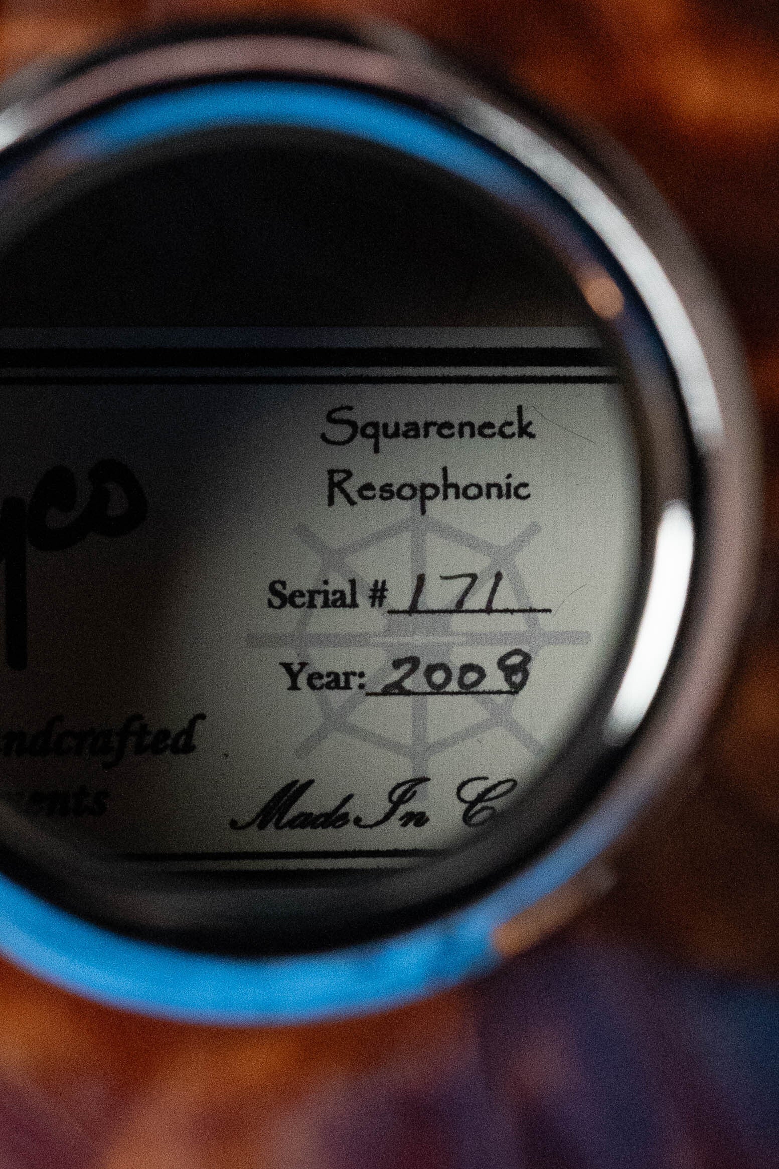 A close-up of a metal label inside a Rayco All Quilted Maple Resonator Acoustic Guitar (#2008), reading: Squareneck Resophonic, Serial #171, Year: 2008, viewed through a circular opening in its quilted maple body.