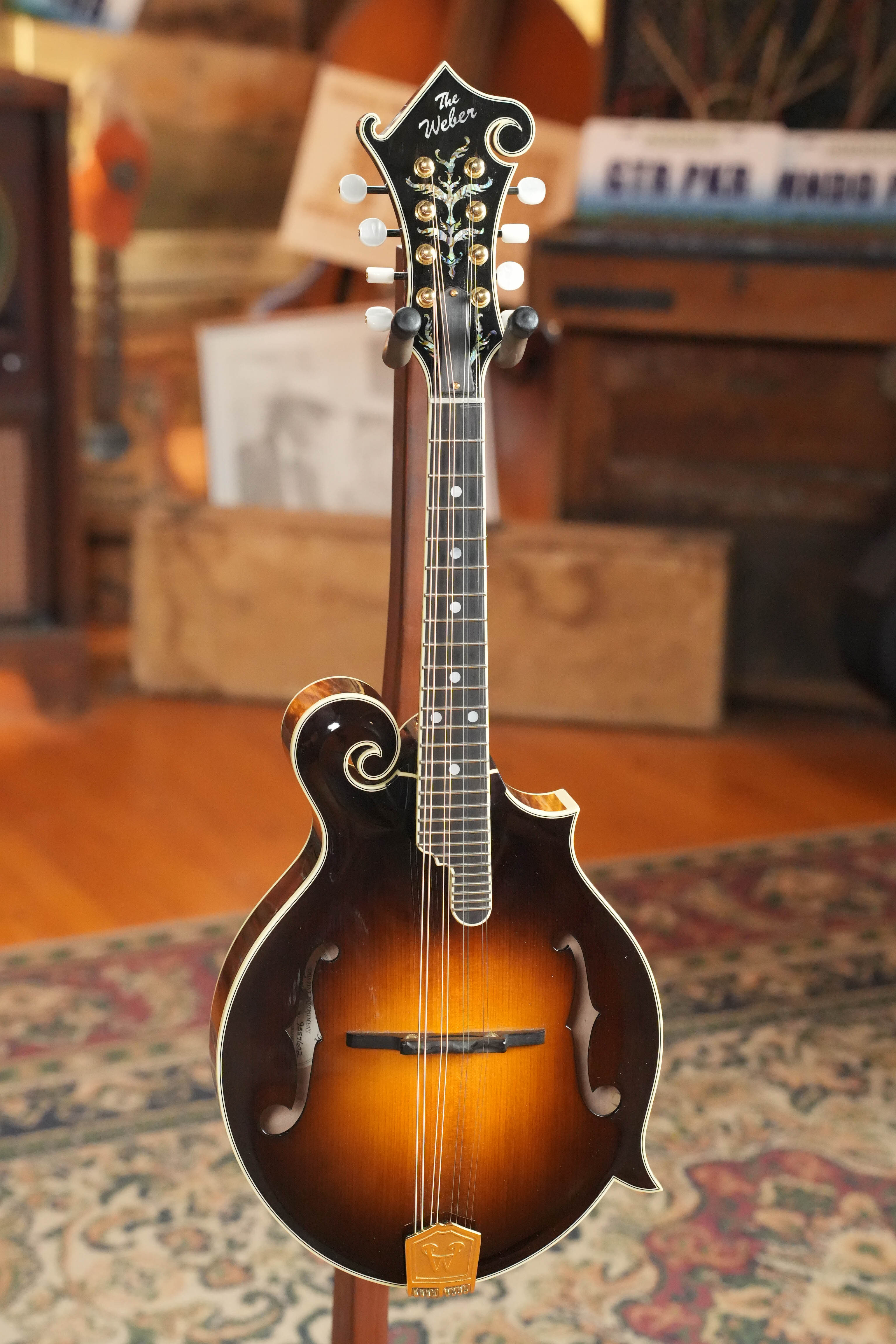 A (Used) 2009 Weber Fern Spruce/Maple F-Style Mandolin #7602 stands upright on a patterned rug in a warmly lit room, showcasing Weber's craftsmanship amid wood furniture and records.