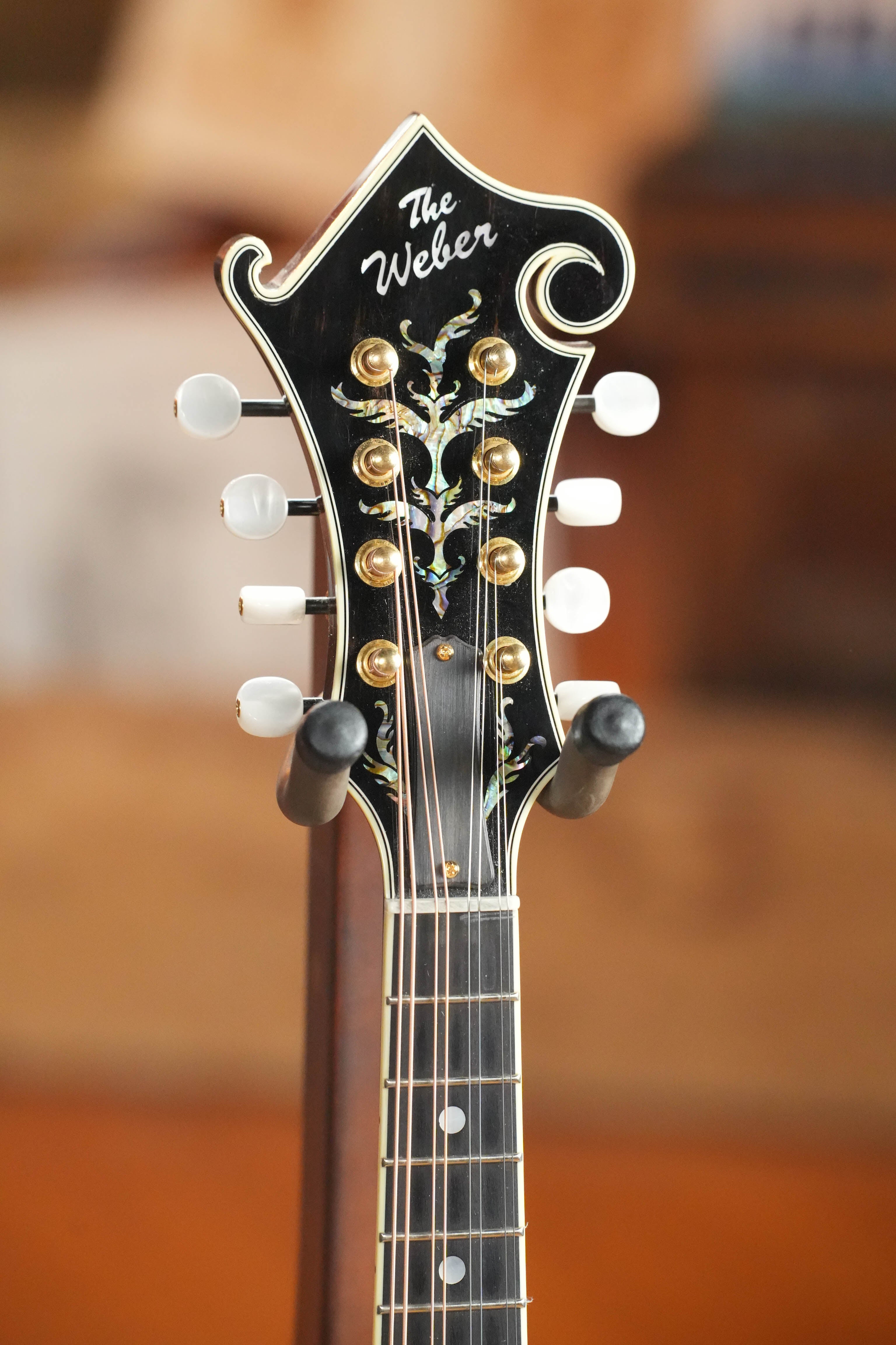 Close-up of the headstock of a (Used) 2009 Weber Fern Spruce/Maple F-Style Mandolin #7602 with gold tuning pegs, white knobs, ornate vine inlay, and The Weber logo—showcasing boutique craftsmanship on a stand.