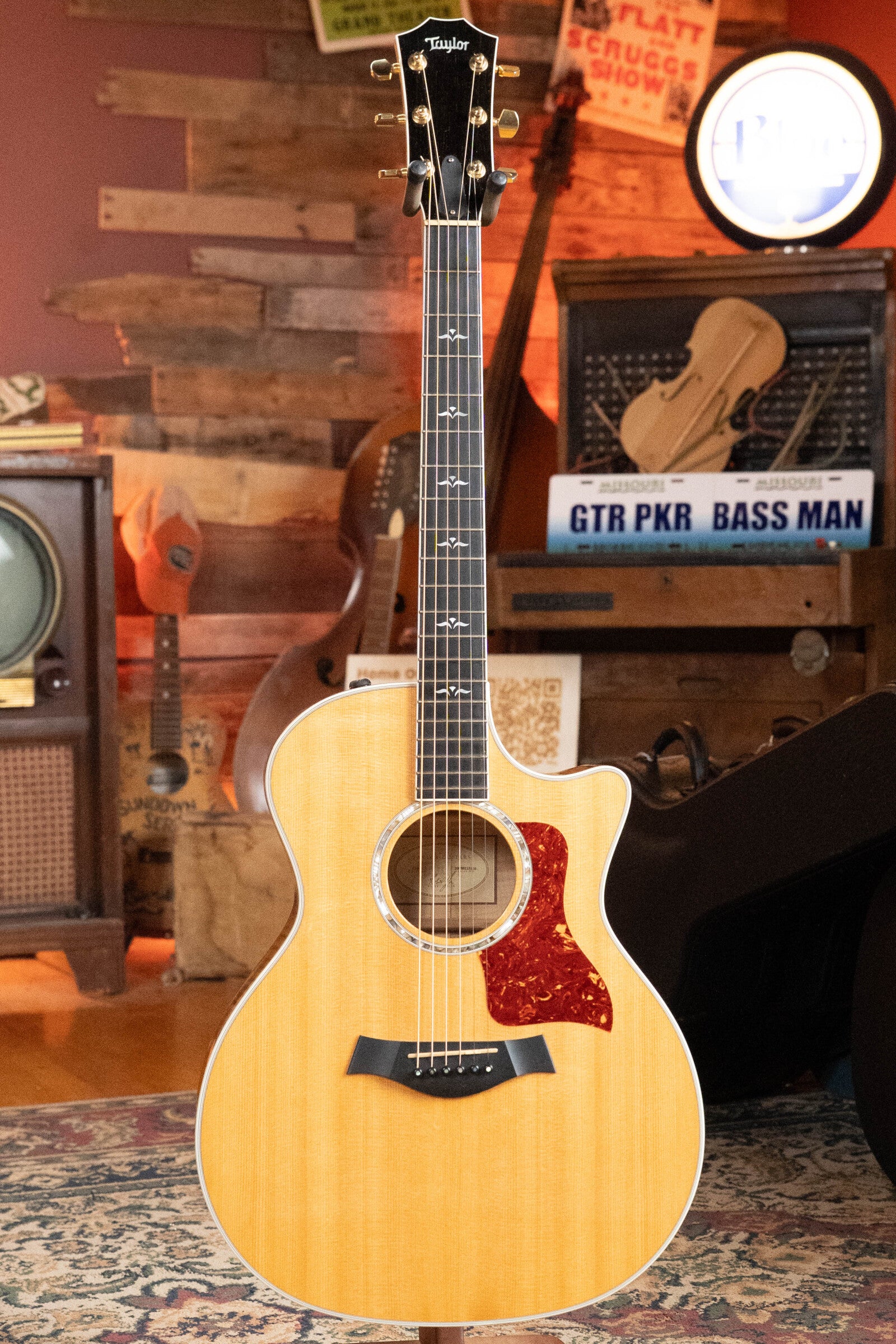 A (Used) 2009 Taylor 614CE Torrefied Sitka/Maple Cutaway Grand Auditorium Guitar w/Pickup by Taylor Guitars stands upright in a cozy room with vintage music gear, including a radio, guitar cases, and music-themed decor.