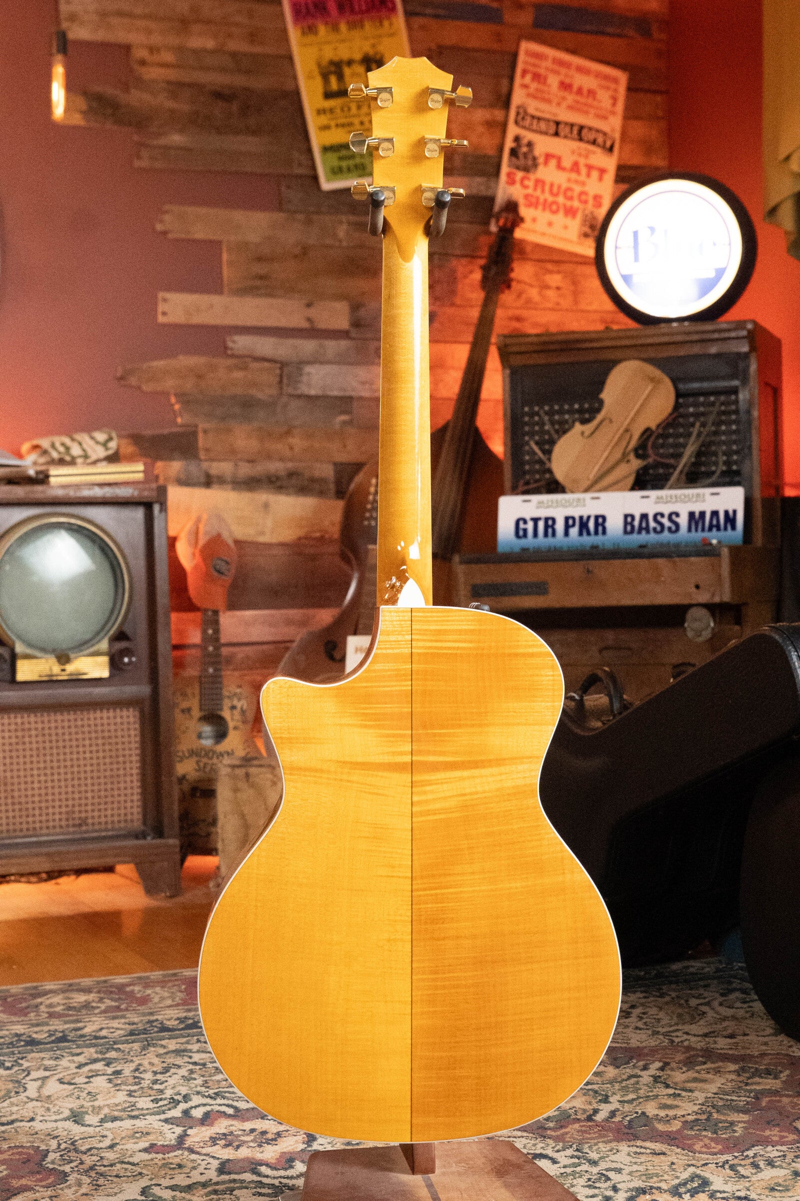 A (Used) 2009 Taylor 614CE Torrefied Sitka/Maple Grand Auditorium guitar with pickup (#5110) by Taylor Guitars is displayed from the back on a rug, against vintage decor featuring a TV, amp, and music posters on a wooden wall.