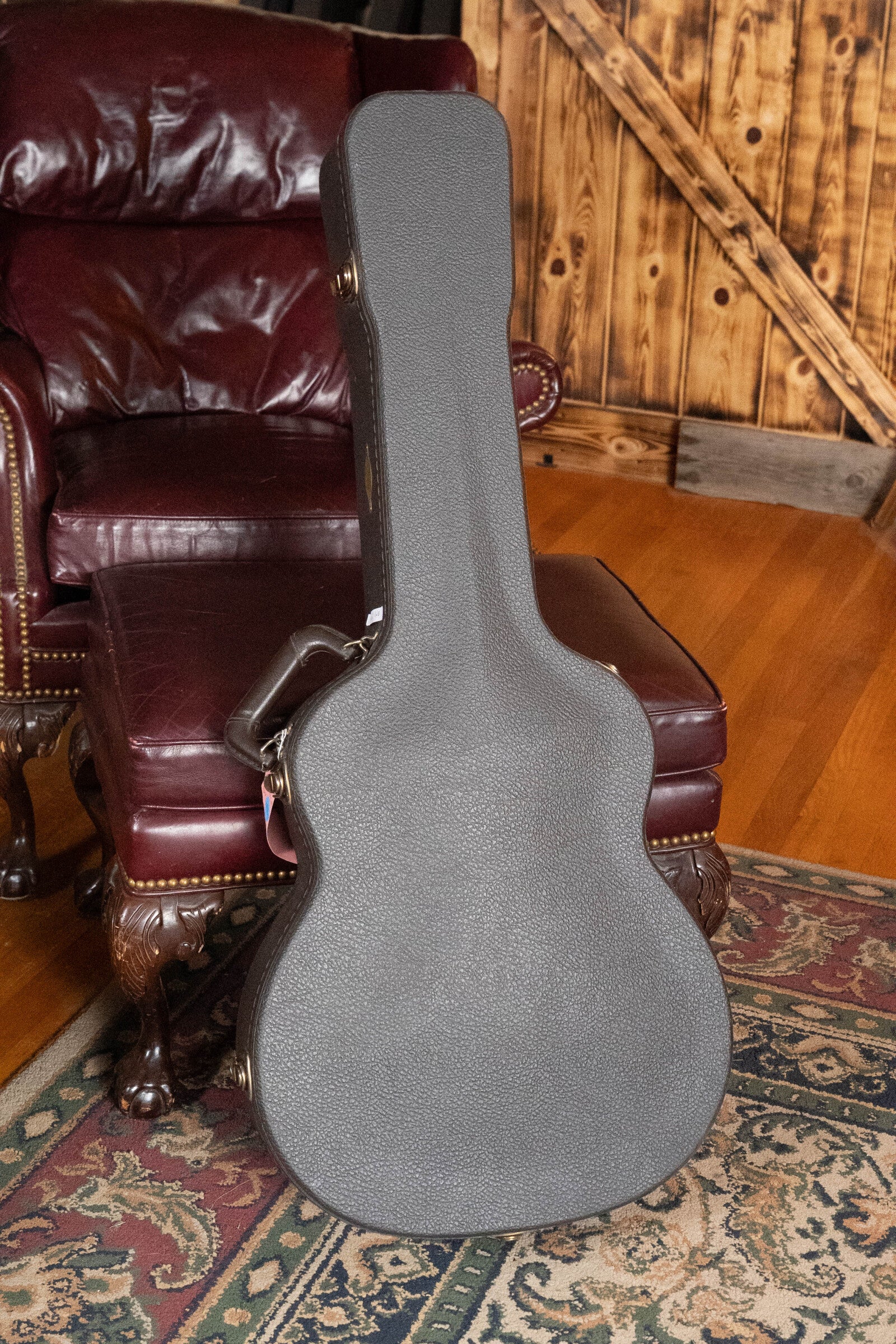 A (Used) 2009 Taylor 614CE Torrefied Sitka/Maple Grand Auditorium Guitar case sits upright on a brown leather ottoman beside a matching chair, with a wooden wall and patterned rug in the background.