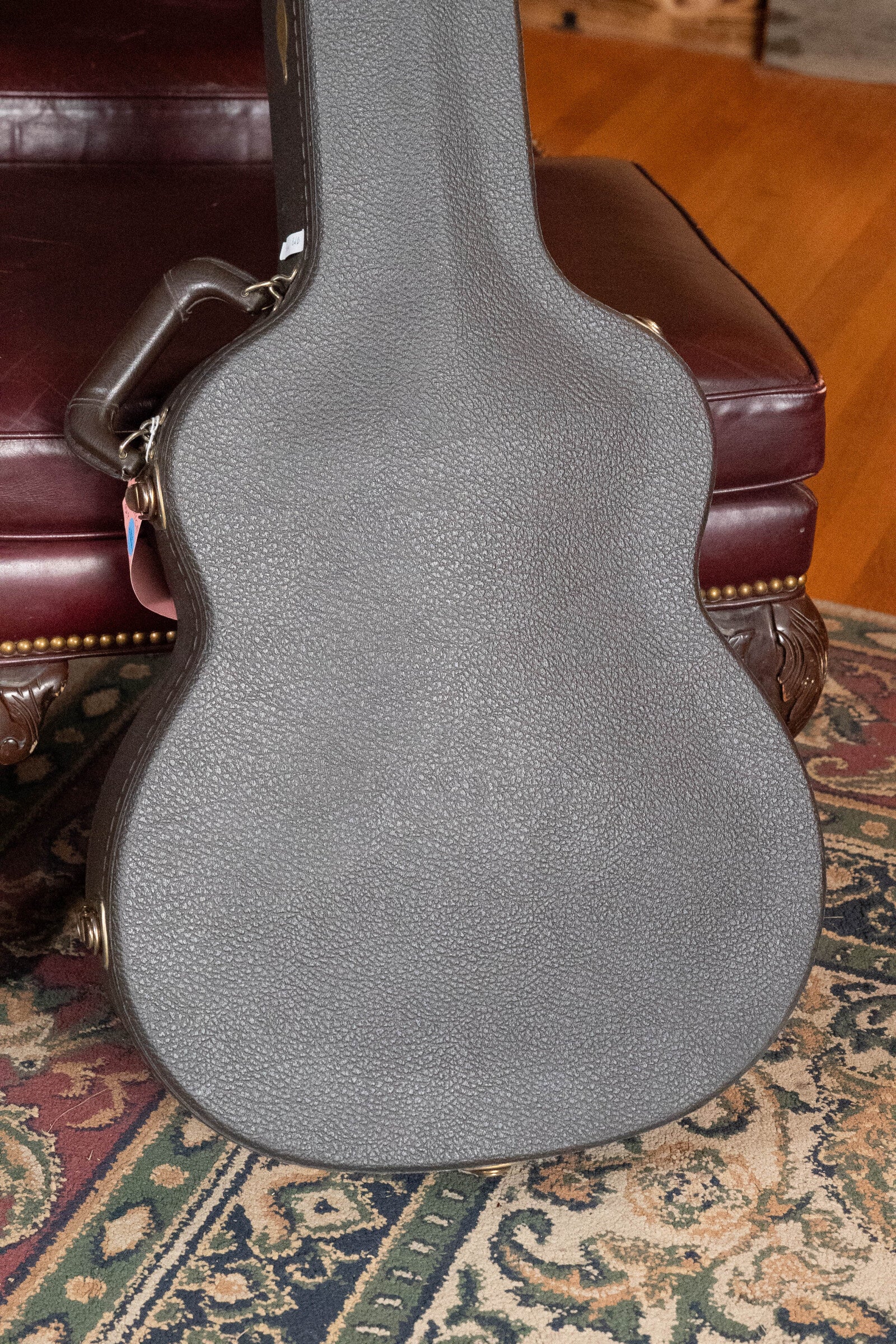 A closed, textured gray case for a (Used) 2009 Taylor 614CE Grand Auditorium guitar by Taylor Guitars stands upright against a maroon leather chair, partially over a patterned rug on a wooden floor.