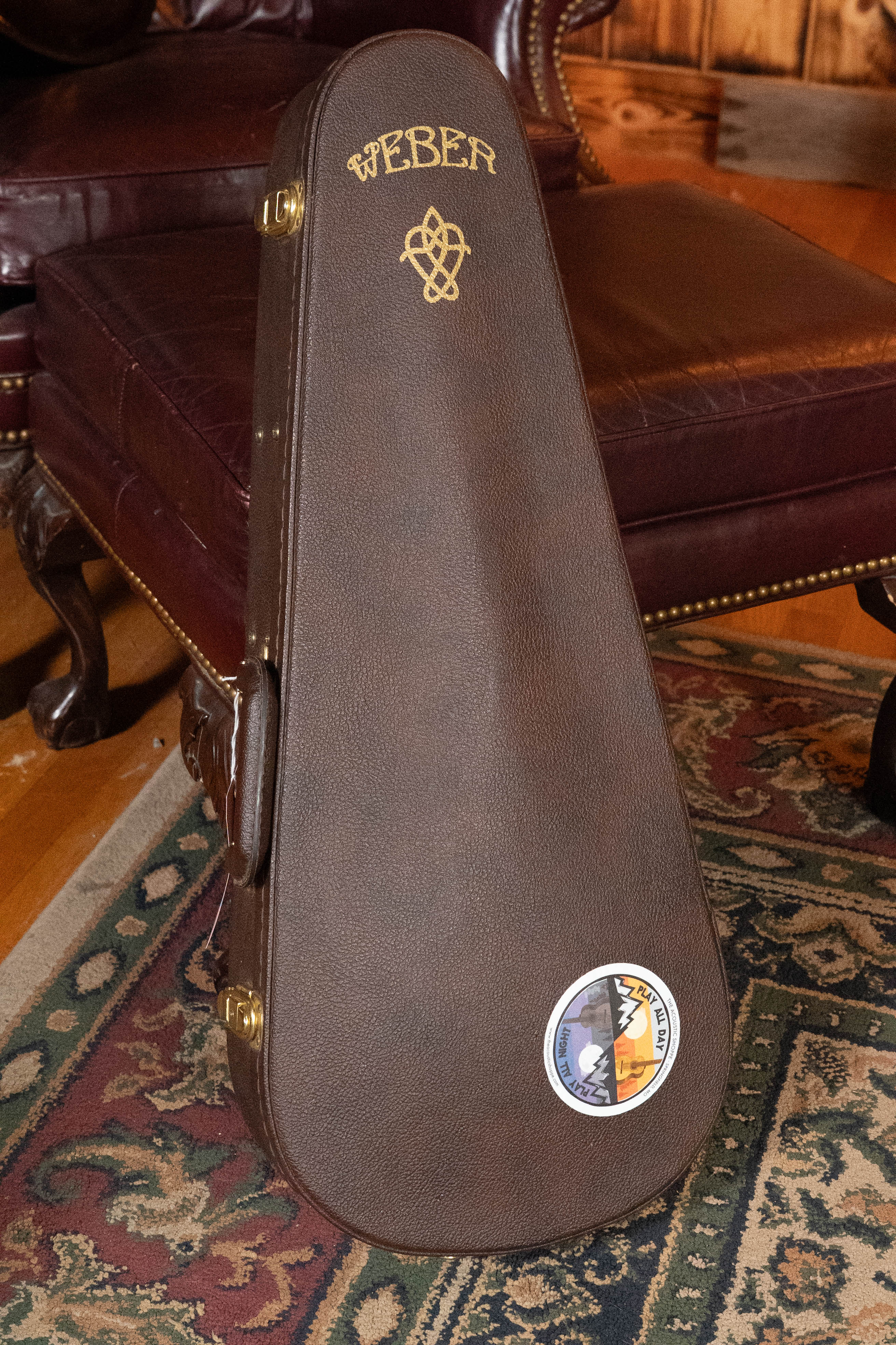 A brown Weber case with gold lettering, a Celtic knot design, gold hardware, and a round sticker—likely for the (Used) 2016 Weber Fern Red Spruce/Flamed Maple A-Style Mandolin #2801—rests against a maroon leather chair.