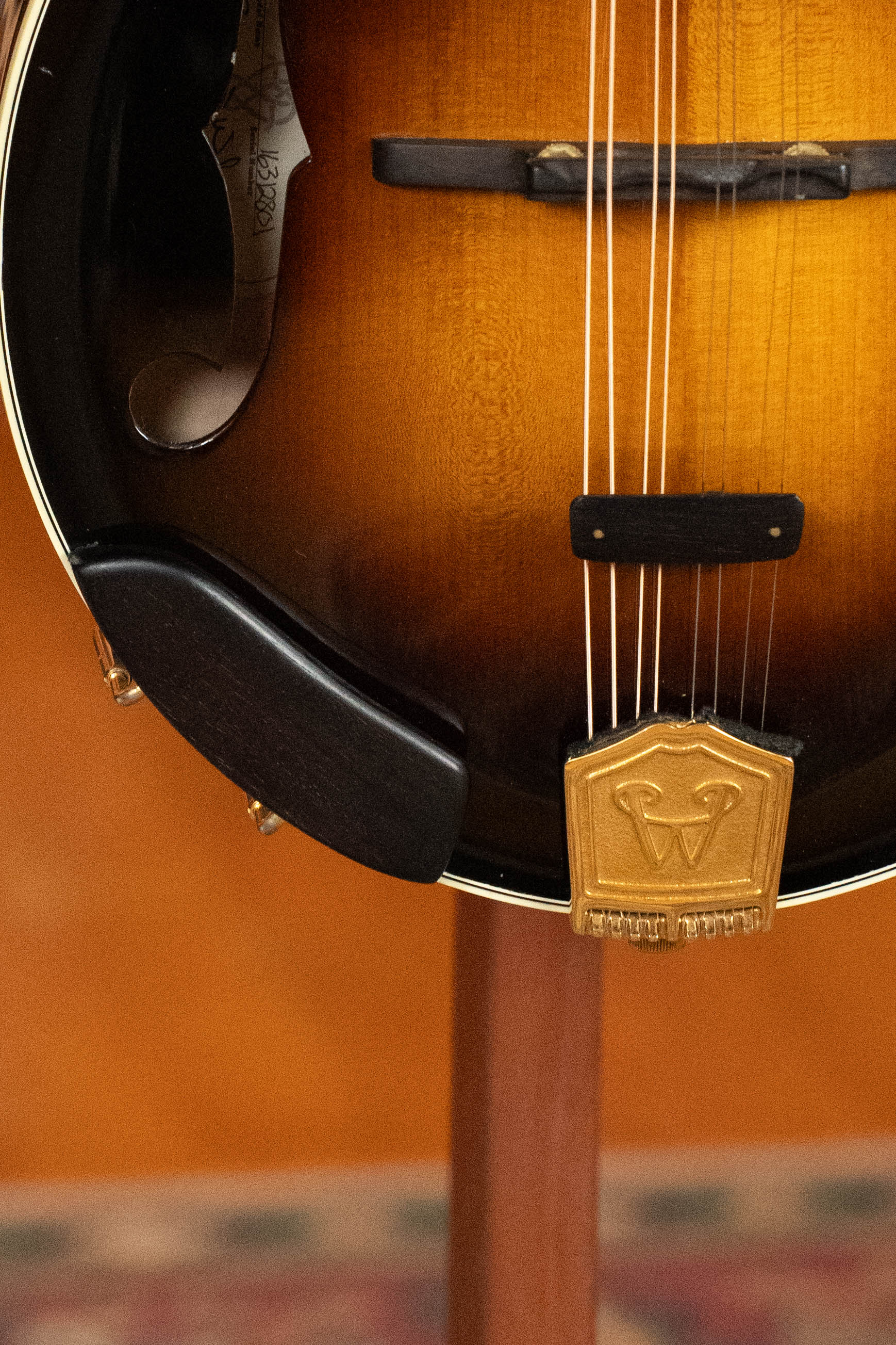 Close-up of the lower half of a (Used) 2016 Weber Fern A-Style Mandolin (#2801), showing its brown sunburst finish, strings, bridge, black chin rest, and gold tailpiece embossed with a W. The warm-toned background is softly blurred.