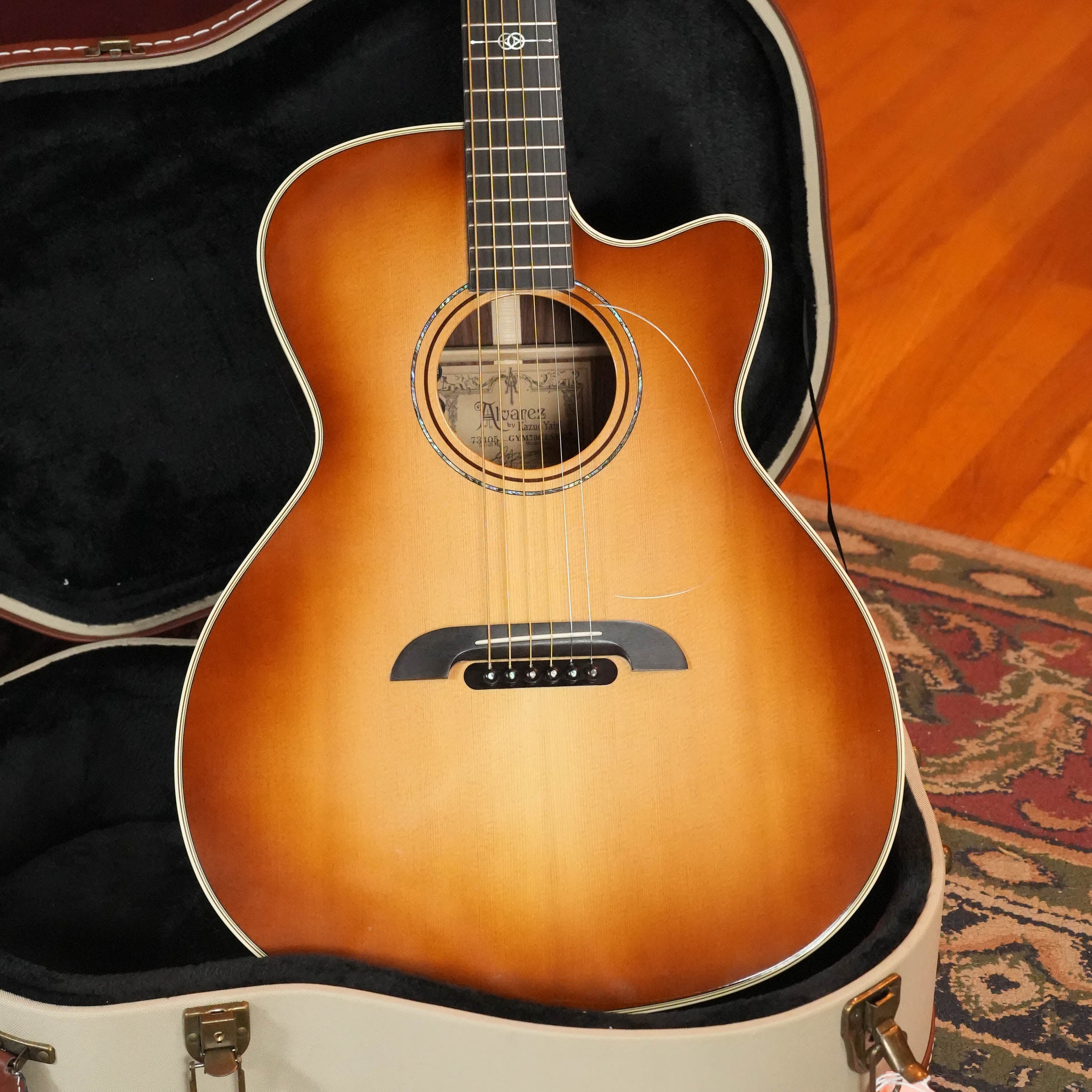A 2018 Alvarez Yairi GYM70CESHB Grand Auditorium acoustic guitar with solid AAA Sitka spruce and East Indian rosewood, from The Acoustic Shoppe, rests in an open hard case on a wooden floor beside a patterned rug.