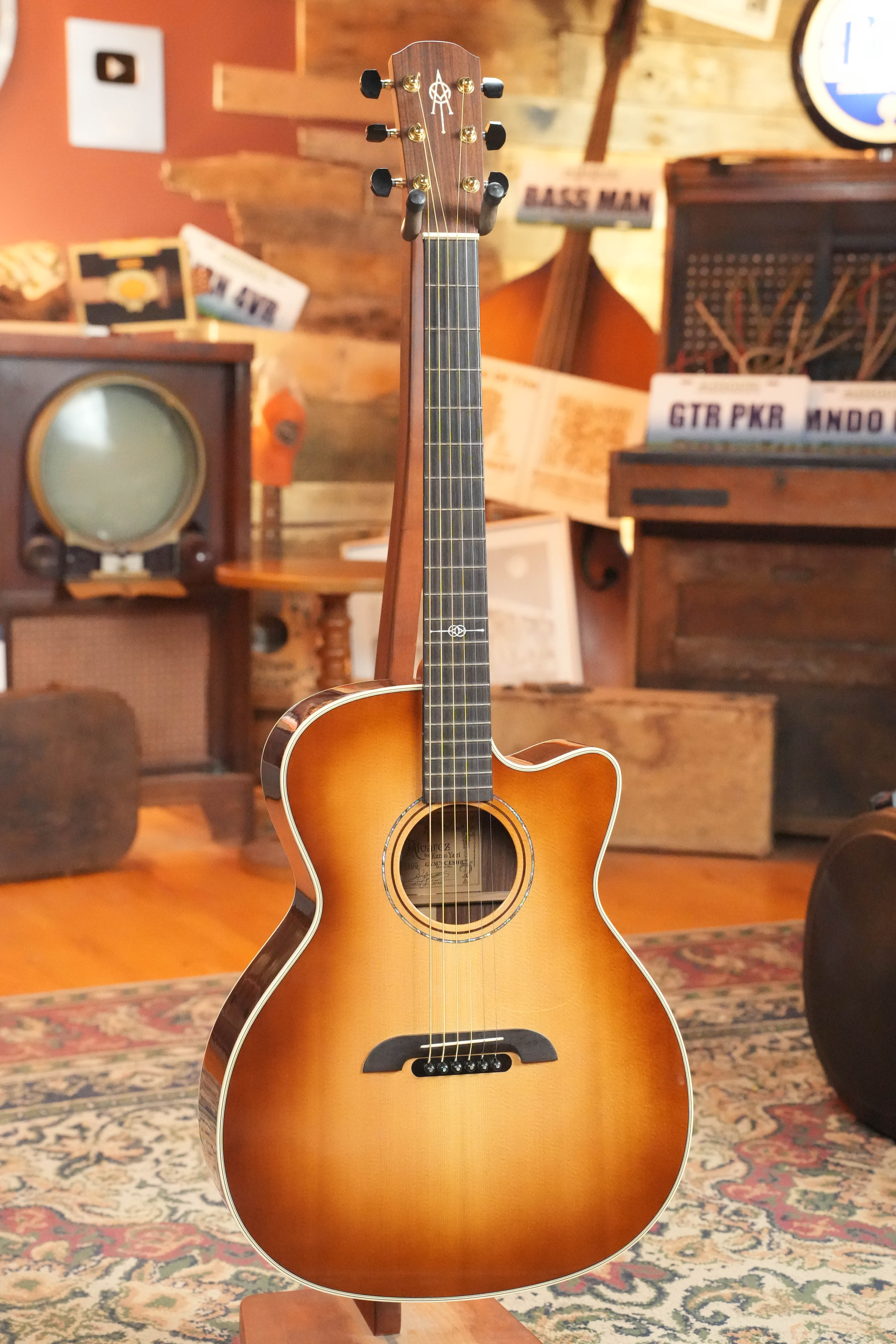 A (Used) 2018 Alvarez Yairi GYM70CESHB Grand Auditorium Acoustic Guitar from The Acoustic Shoppe stands on a wooden floor in a cozy, vintage-style room filled with retro decor and musical instruments.