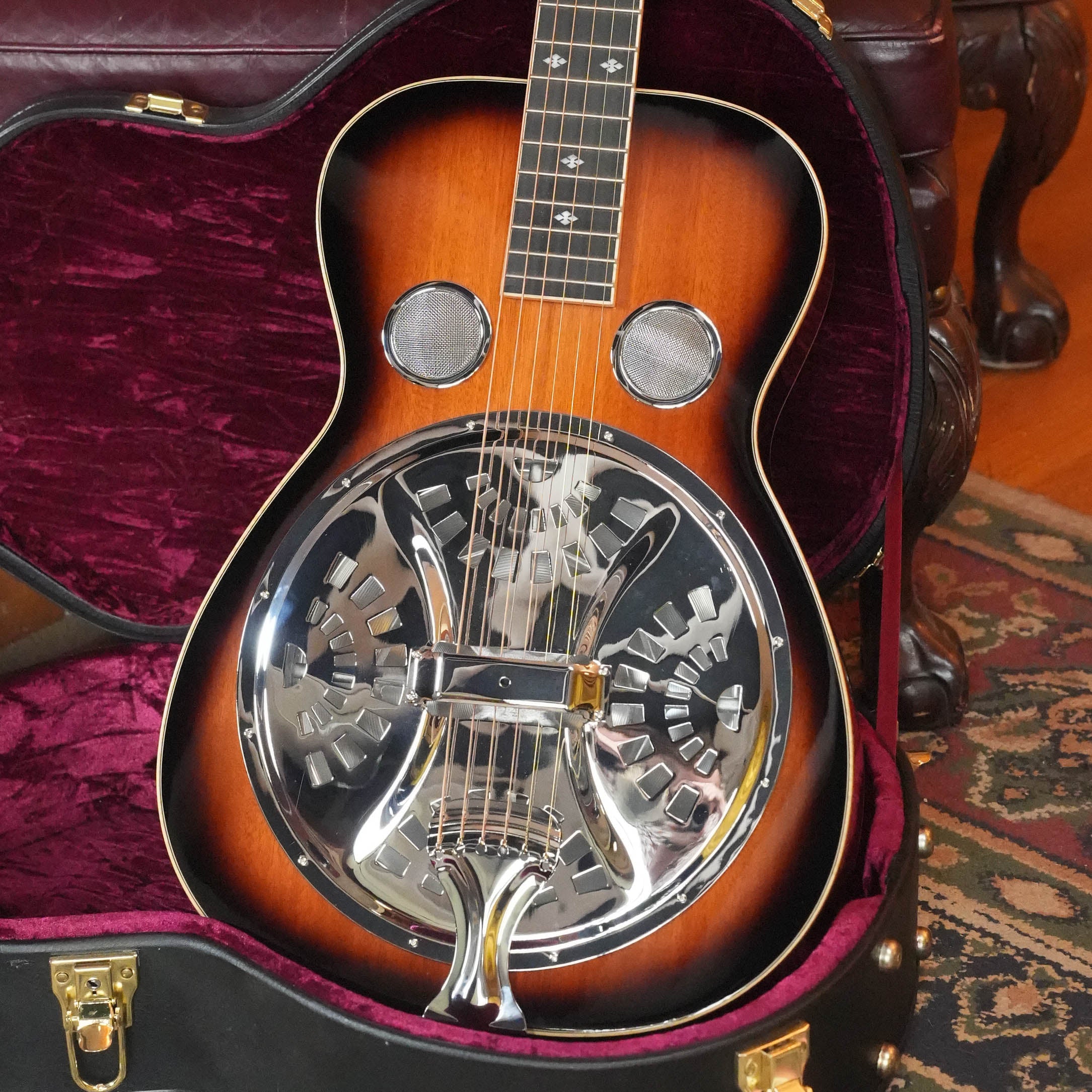 A (Used) 2022 Gold Tone PBS-D Paul Beard Deluxe Solid Mahogany Square-Neck Resonator Guitar #8165 with a sunburst finish sits in a plush, red-lined case, featuring a shiny metal resonator cone; part of a rug and wood floor are visible.