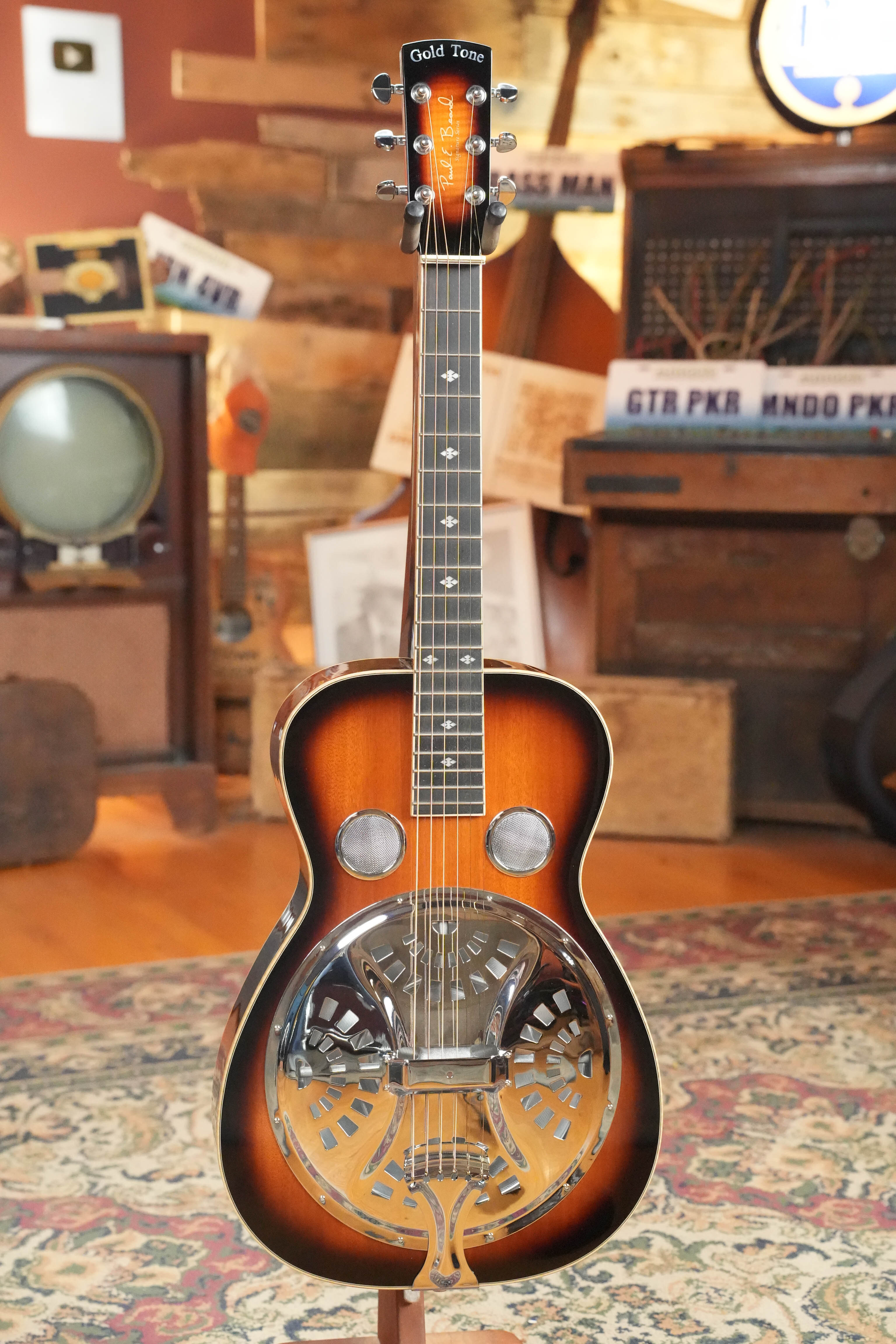 A (Used) 2022 Gold Tone PBS-D Paul Beard Deluxe Solid Mahogany Square-Neck Resonator Guitar #8165 stands upright on a patterned rug in a vintage-themed room with an old TV, suitcases, and retro signs in the background.