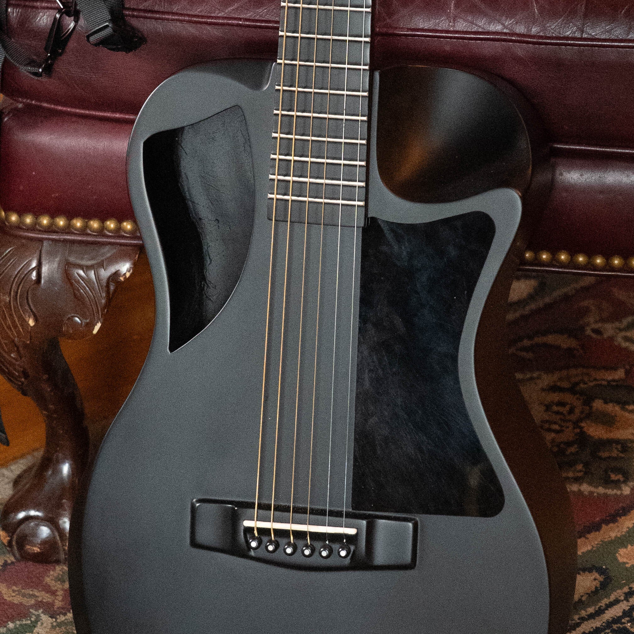 A close-up of a matte black 2022 Journey Instruments OF660M all carbon fiber compact travel guitar with JourneyTek-3 pickup, shown resting against a maroon leather chair on a patterned rug.