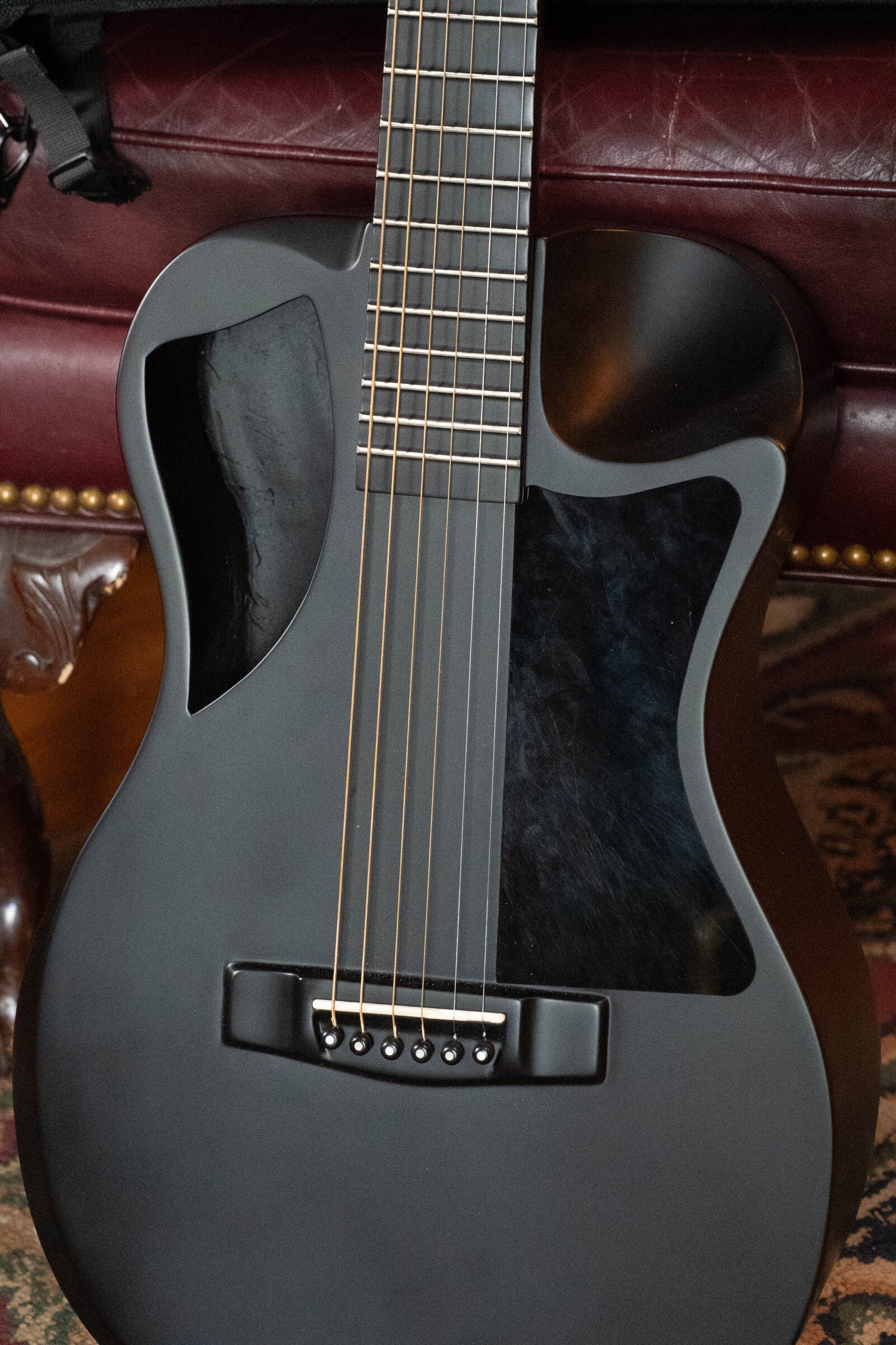 A close-up of a matte black 2022 Journey Instruments OF660M all carbon fiber compact travel guitar with a minimalist design and unique black pickguards, resting on a maroon leather couch with brass studs.