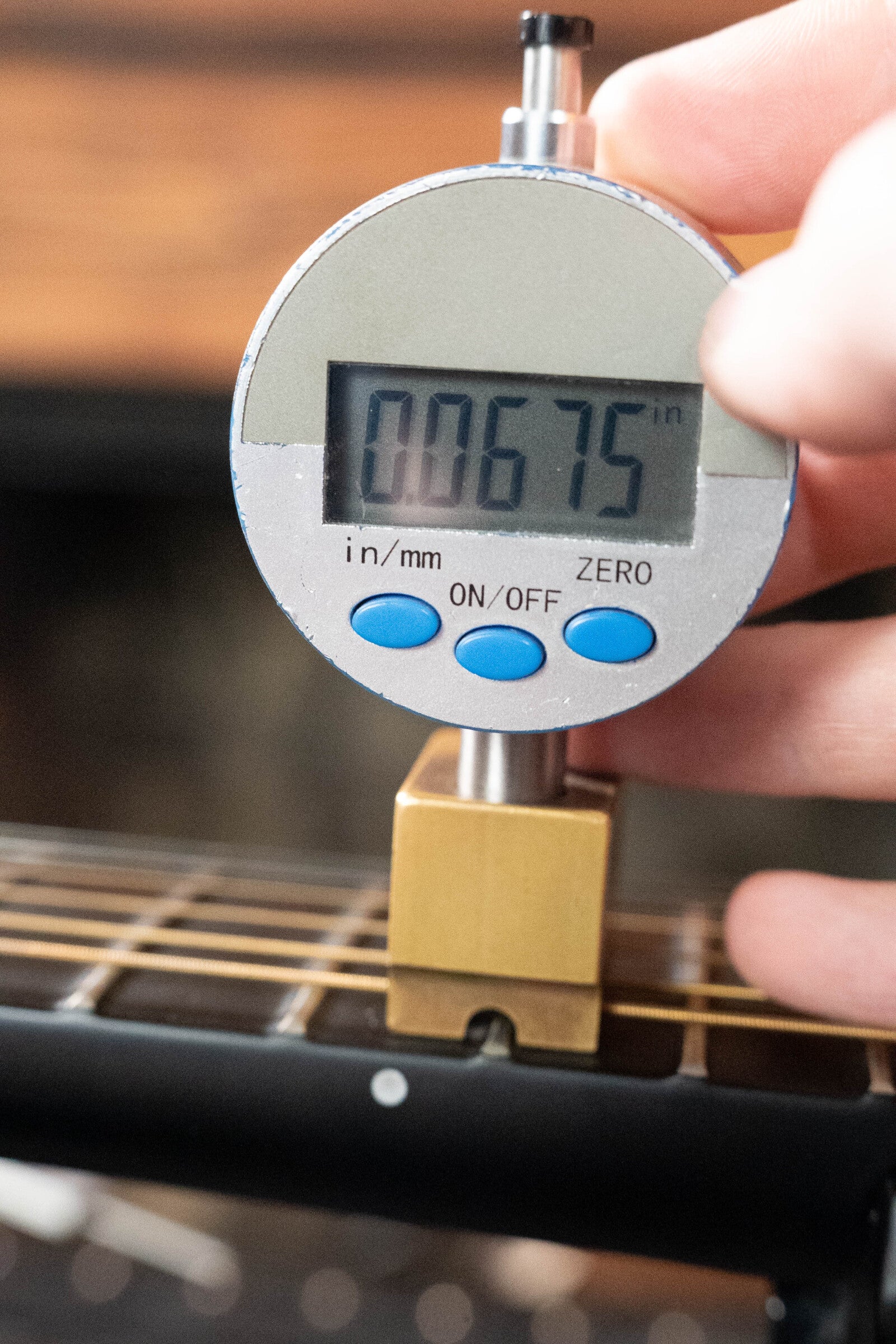 A digital gauge shows a 0.0675-inch string height above the fretboard of a (Used) 2022 Journey Instruments OF660M All Carbon Fiber Compact OO Travel Guitar with JouorneyTek-3 Pickup, held over its sleek neck.