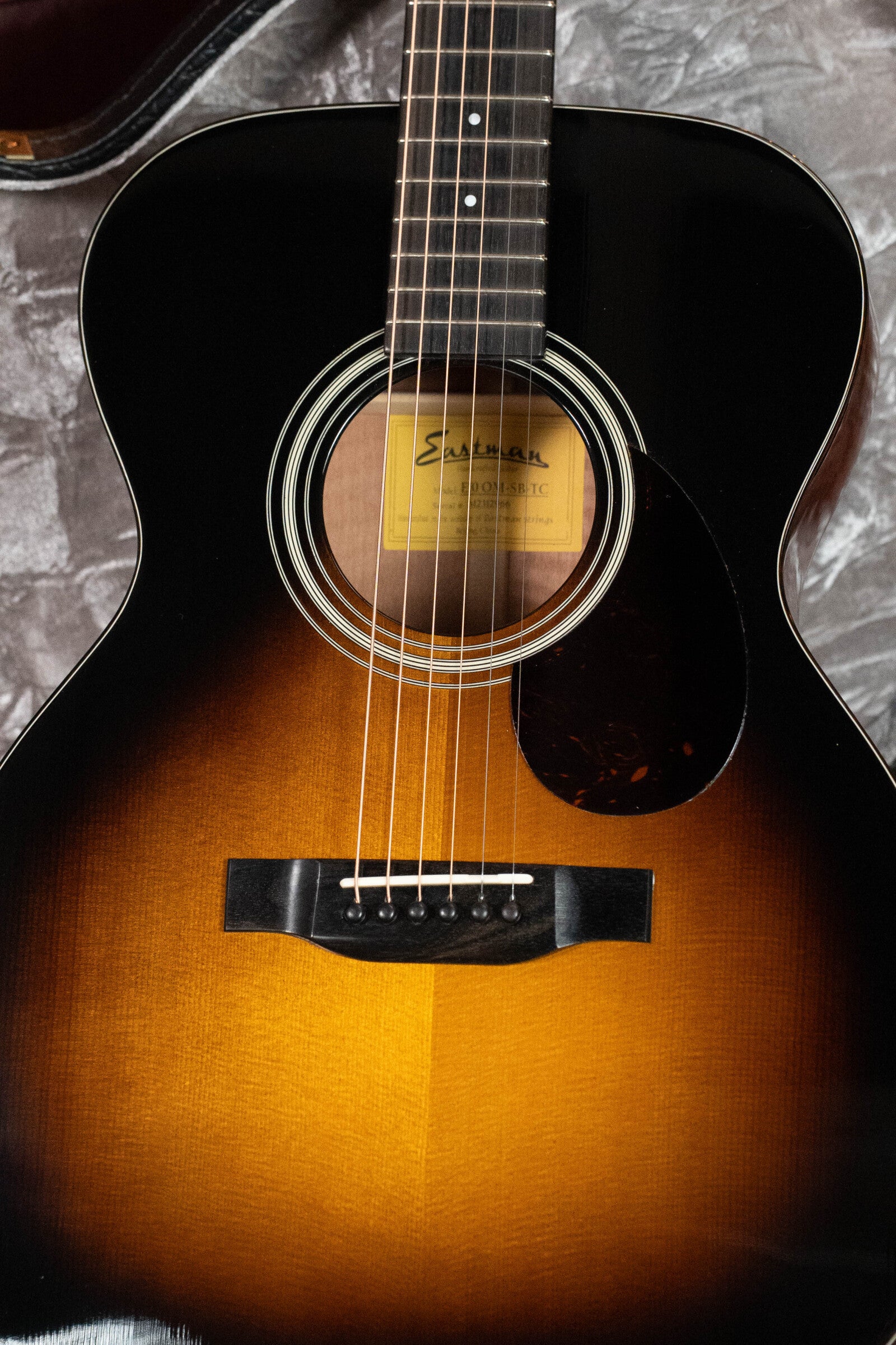 A close-up of a (Used) 2023 Eastman Guitars E10OM-TC-SB Orchestra Model shows its sunburst Thermo-Cured Adirondack Spruce top, mahogany body, strings, frets, sound hole, and label on a textured gray surface.