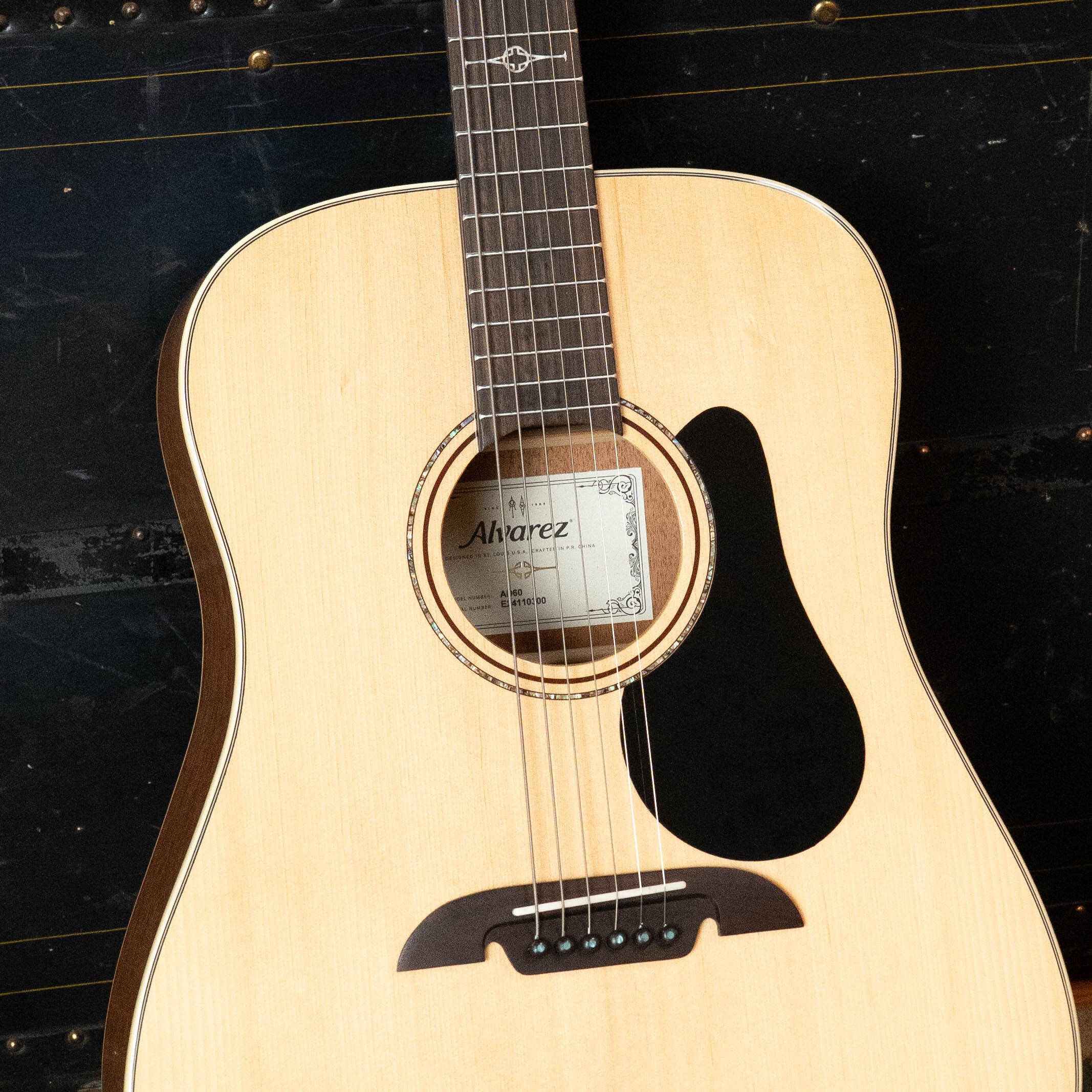 Close-up of a (Used) 2024 Alvarez AD60 Sitka/Mahogany Dreadnought Acoustic Guitar #0300 featuring a light wood top and dark pickguard; Alvarez is visible on the label inside the soundhole against a dark, textured background.