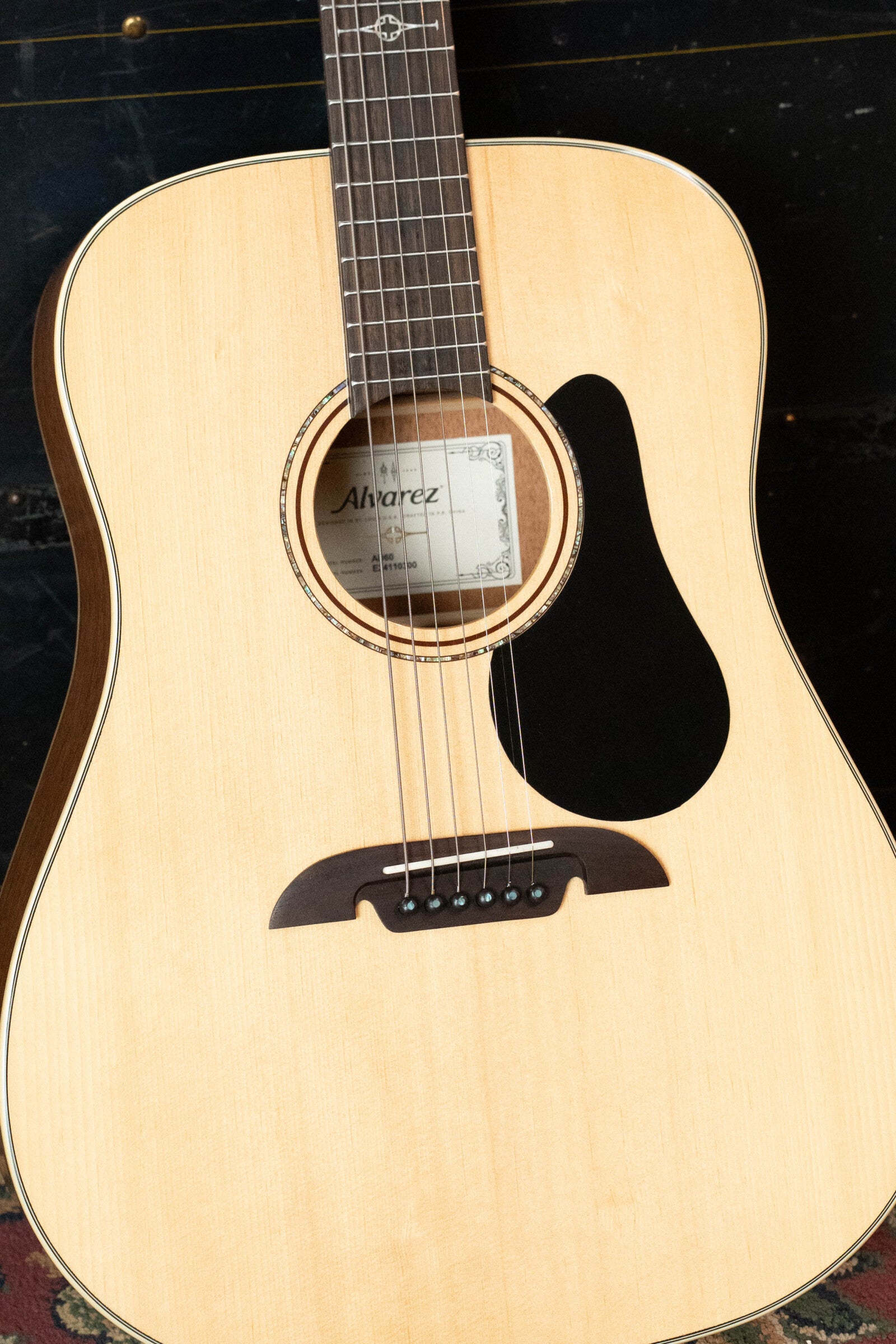 A close-up of a (used) 2024 Alvarez AD60 Sitka/Mahogany Dreadnought Acoustic Guitar #0300 shows its light wood body, visible strings, and black pickguard. The Alvarez label is seen through the sound hole as it stands upright on a dark surface.