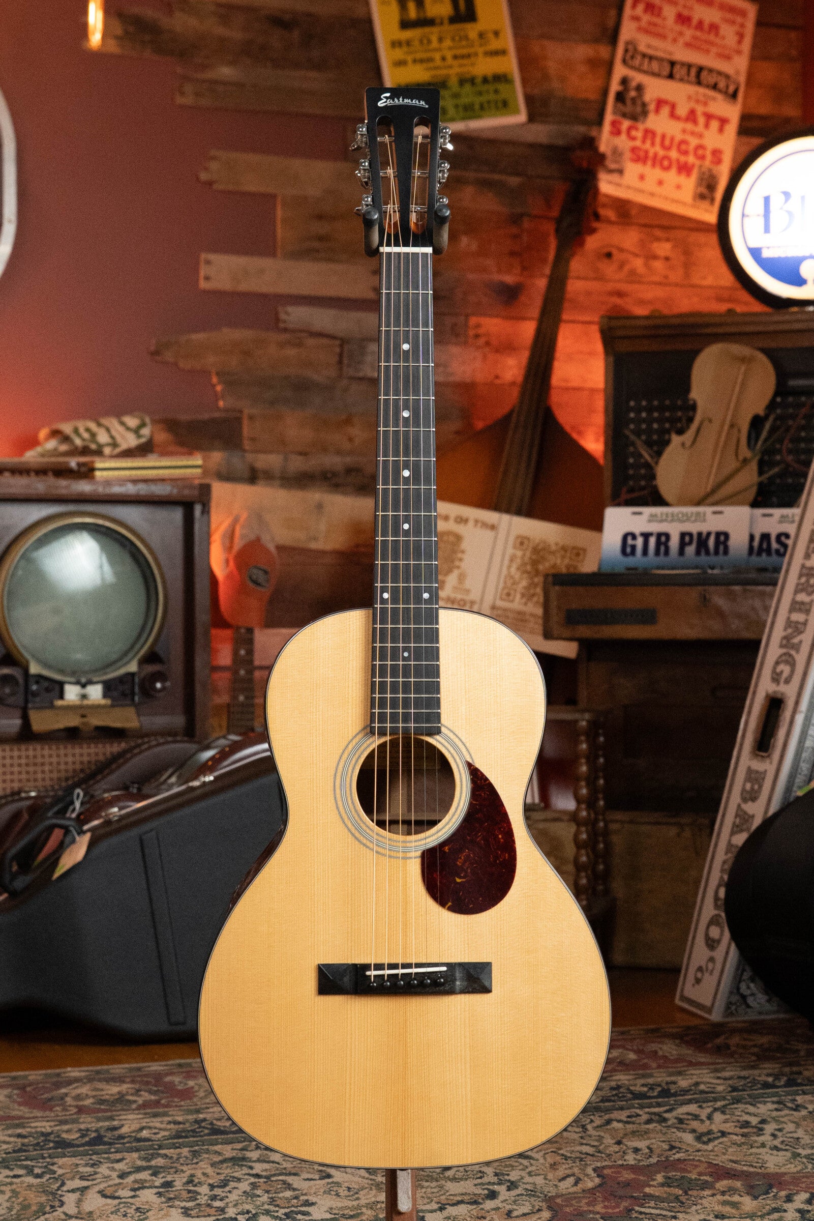 A (Used) 2024 Eastman Guitars E10OO-TC Thermo-Cure Adirondack/Mahogany Acoustic Guitar #0268 with a light wood body and dark neck stands upright in a vintage room with wooden walls, old radio, posters, and musical equipment.