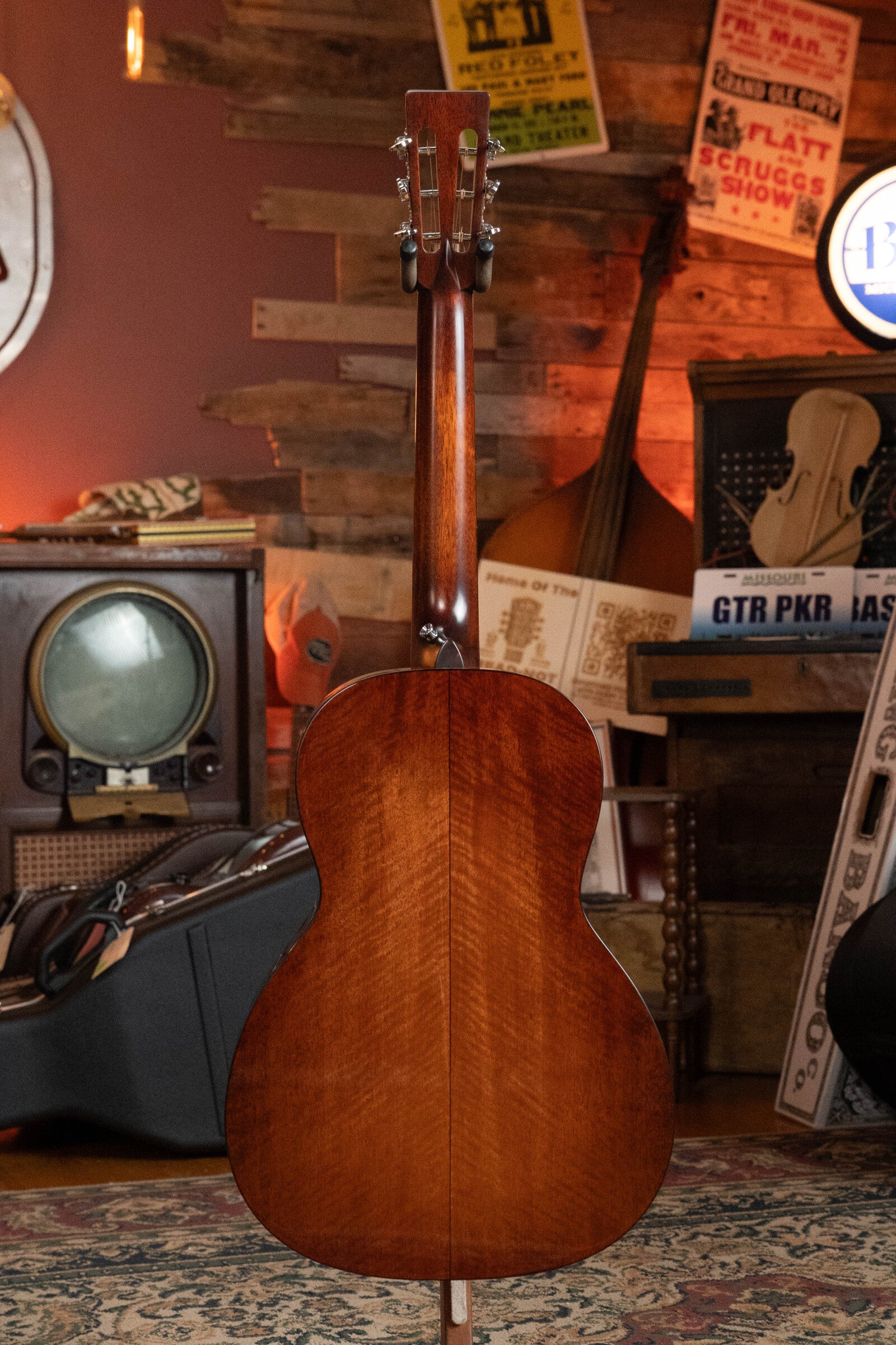 A (Used) 2024 Eastman Guitars E10OO-TC Thermo-Cure Adirondack/Mahogany Acoustic Guitar #0268 is displayed upright from the back in a cozy room with vintage music posters, an old TV, and various musical instruments and equipment.