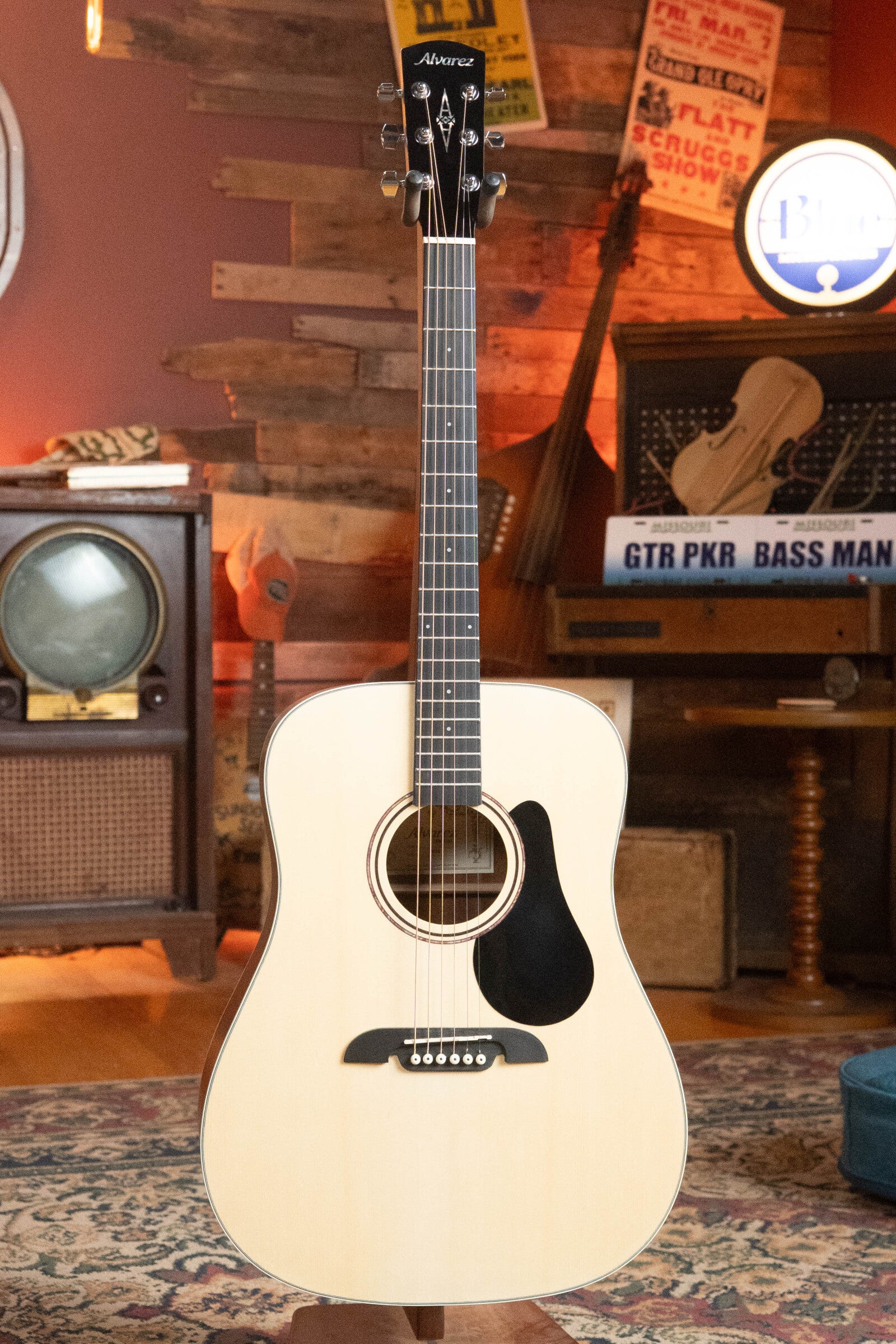 An Alvarez (Used) 2025 RD-26 Spruce/Mahogany Dreadnought Acoustic Guitar #0161 stands upright in a cozy vintage room with wood paneling, a retro TV, rug, and musical decor in the background.