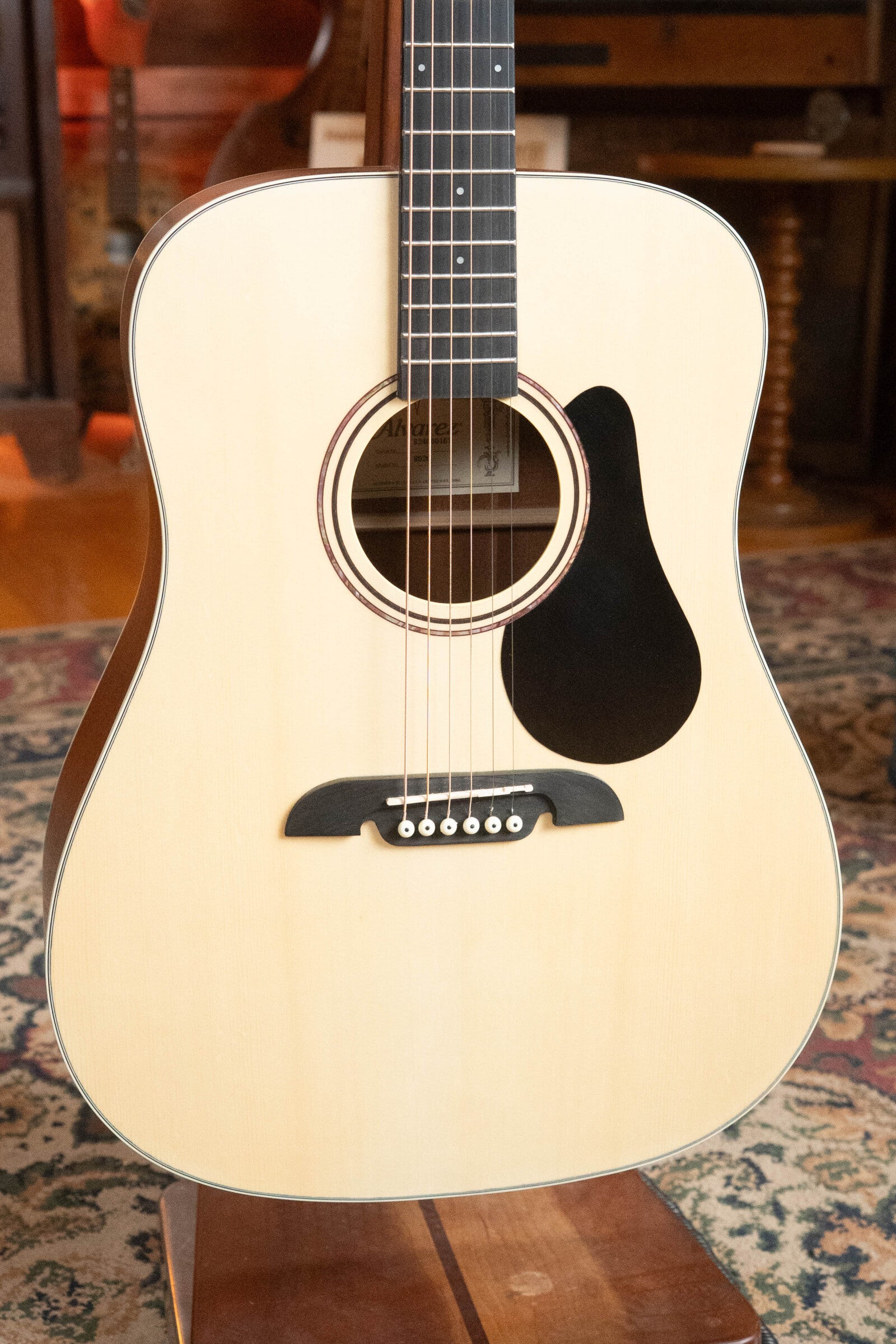 A close-up of a (Used) 2025 Alvarez RD-26 Spruce/Mahogany Dreadnought Acoustic Guitar #0161 displayed upright on a wooden stand, featuring a light spruce top, dark fretboard, and black pickguard on a patterned carpet.