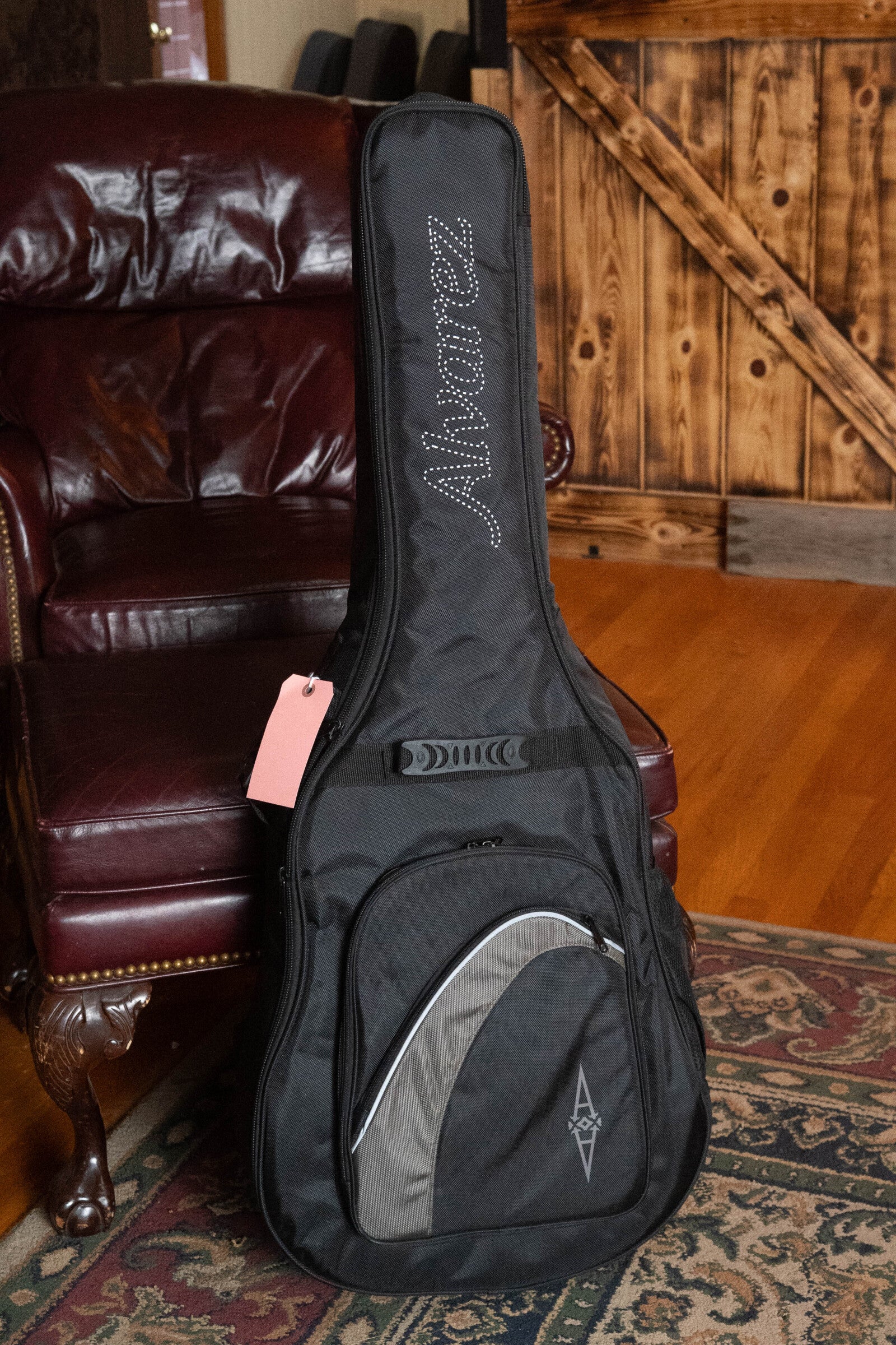 A black Alvarez gig bag stands upright on a patterned rug beside a leather armchair and wooden sliding door, holding the (Used) 2025 Alvarez RD-26 Spruce/Mahogany Dreadnought Acoustic Guitar #0161. A pink tag adds flair.