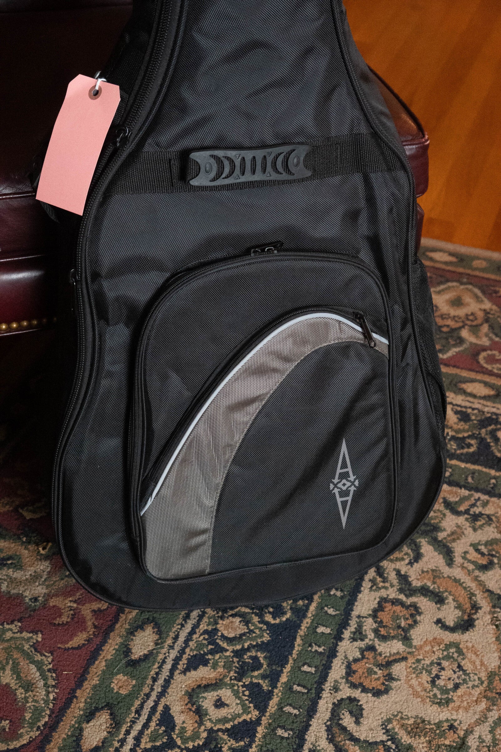 An Alvarez RD-26 Spruce/Mahogany Dreadnought Acoustic Guitar (#0161, used) case with a front pocket, zipper, and pink tag sits on a patterned rug beside a dark red leather chair. Ideal for dreadnought-sized guitars.