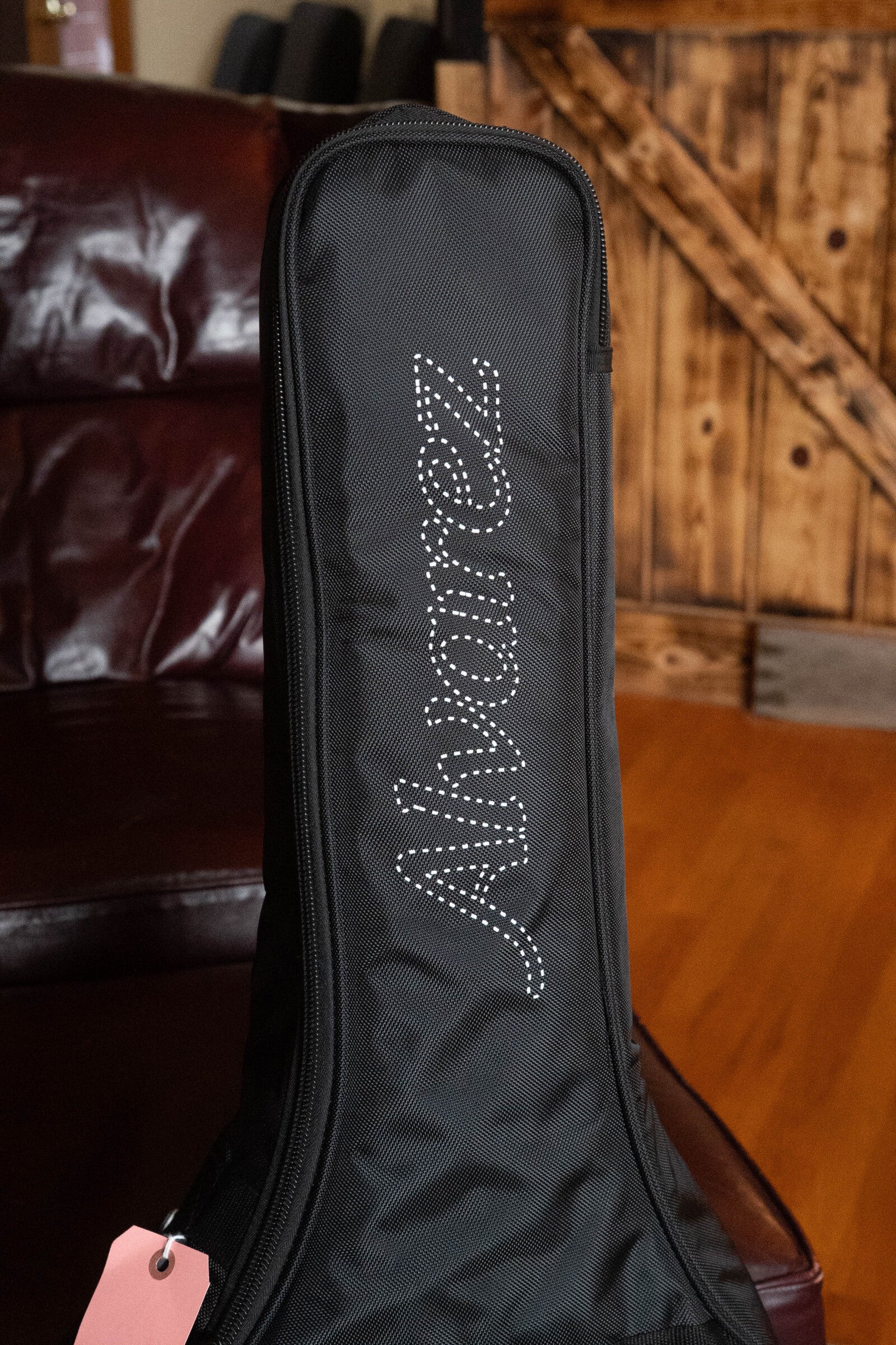 A black Alvarez guitar case with a white logo sits near a brown leather couch and wooden door. A pink tag hangs from the zipper—ideal for your (Used) 2025 Alvarez RD-26 Spruce/Mahogany Dreadnought Acoustic Guitar.