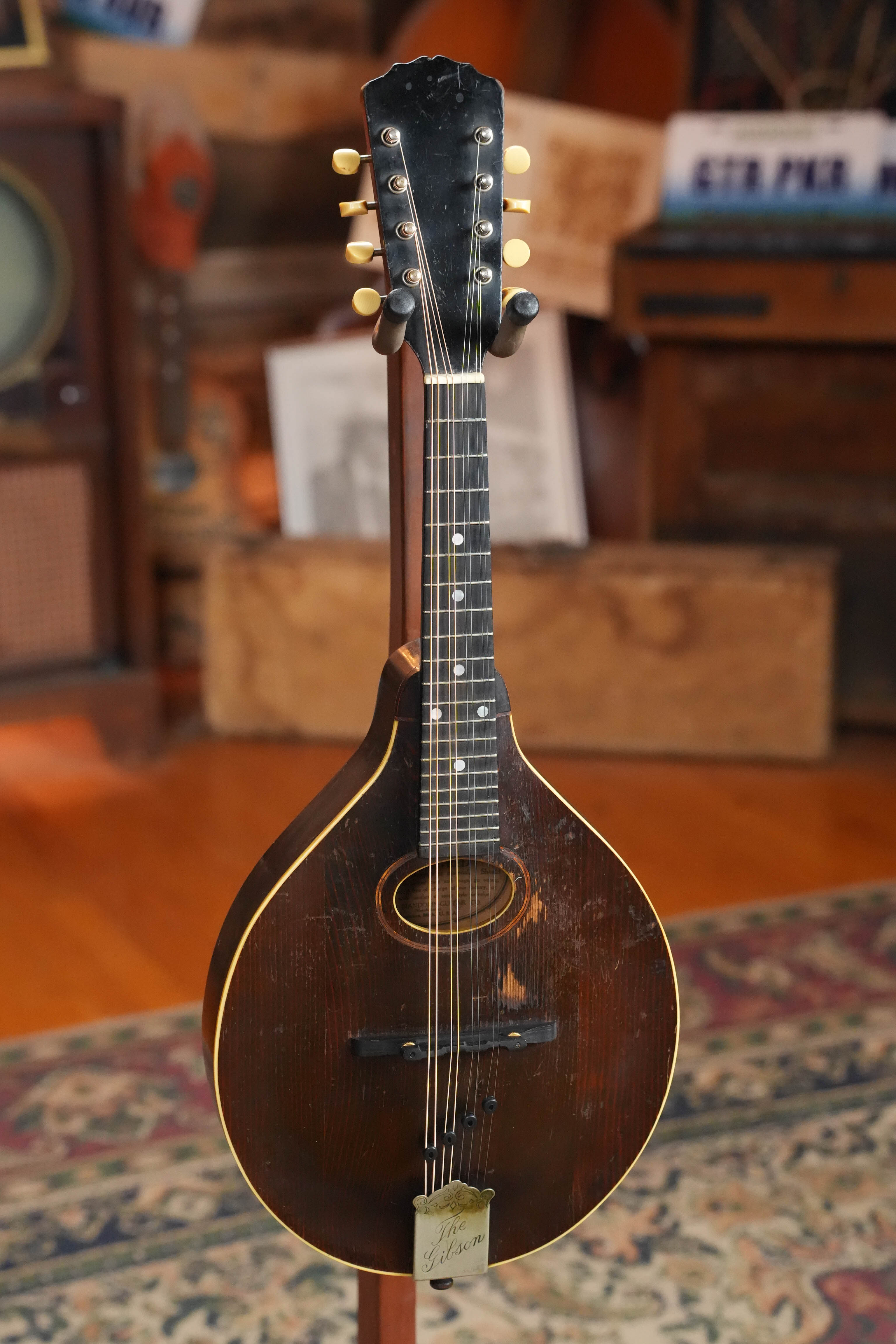 A (Used) Vintage 1920 Gibson Spruce/Birch Oval-Hole Teardrop A-Style Mandolin #9066 by Gibson, featuring eight tuning pegs, stands on a wooden stand in a cozy room with a patterned rug, wood furniture, and books in the background.