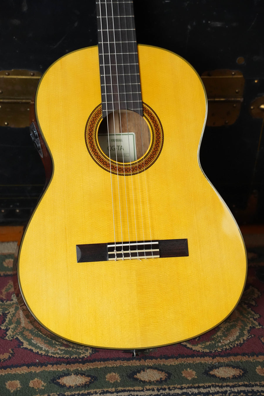 A Yamaha CG-TA Spruce/Ovangkol TransAcoustic Nylon String Acoustic Guitar (#0018) with a solid spruce top stands upright on a patterned rug, with part of a black trunk visible in the background.
