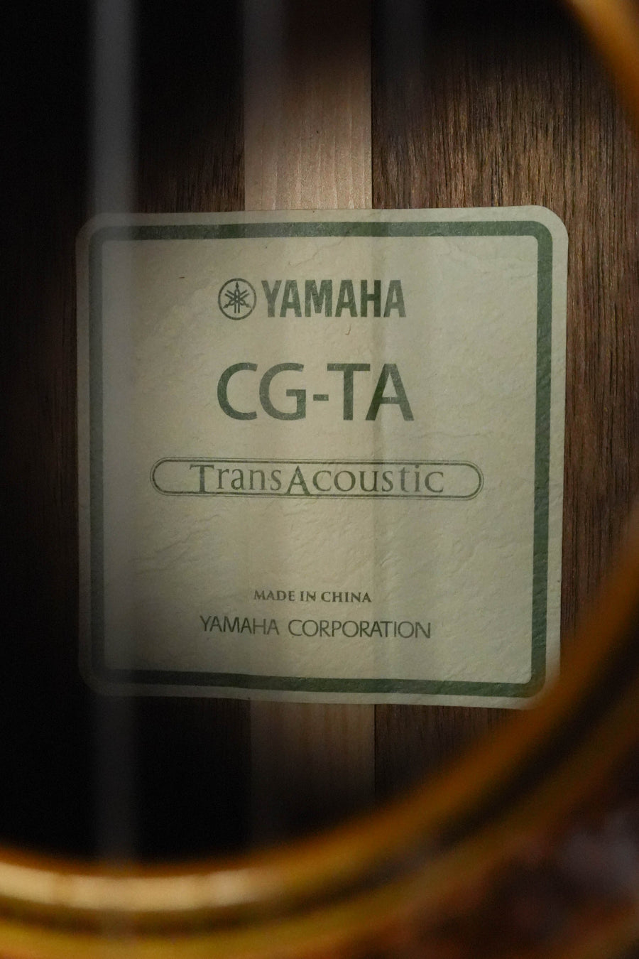 Close-up of a label inside a Yamaha CG-TA Spruce/Ovangkol Transacoustic Nylon String Acoustic Guitar #0018, framed by the sound hole. The label shows the model and reveals it's made by Yamaha.