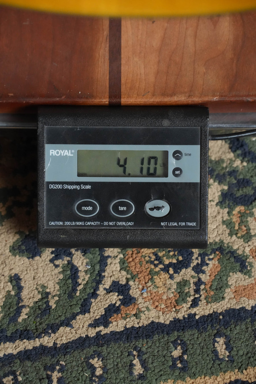 A digital shipping scale on patterned carpet shows a weight of 4.10 lbs, perfect for safely shipping your Yamaha CG-TA Spruce/Ovangkol TransAcoustic Nylon String Acoustic Guitar #0018 by Yamaha.