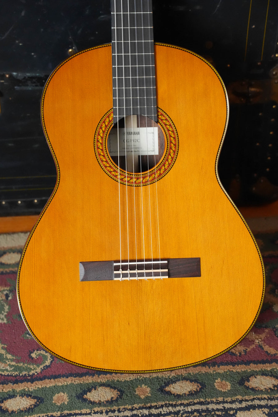 A Yamaha CG192C Cedar/Rosewood Classical Acoustic Guitar #0188 by Yamaha stands upright on a patterned rug, showcasing its cedar top, wooden finish, visible soundhole rosette, and part of the neck.