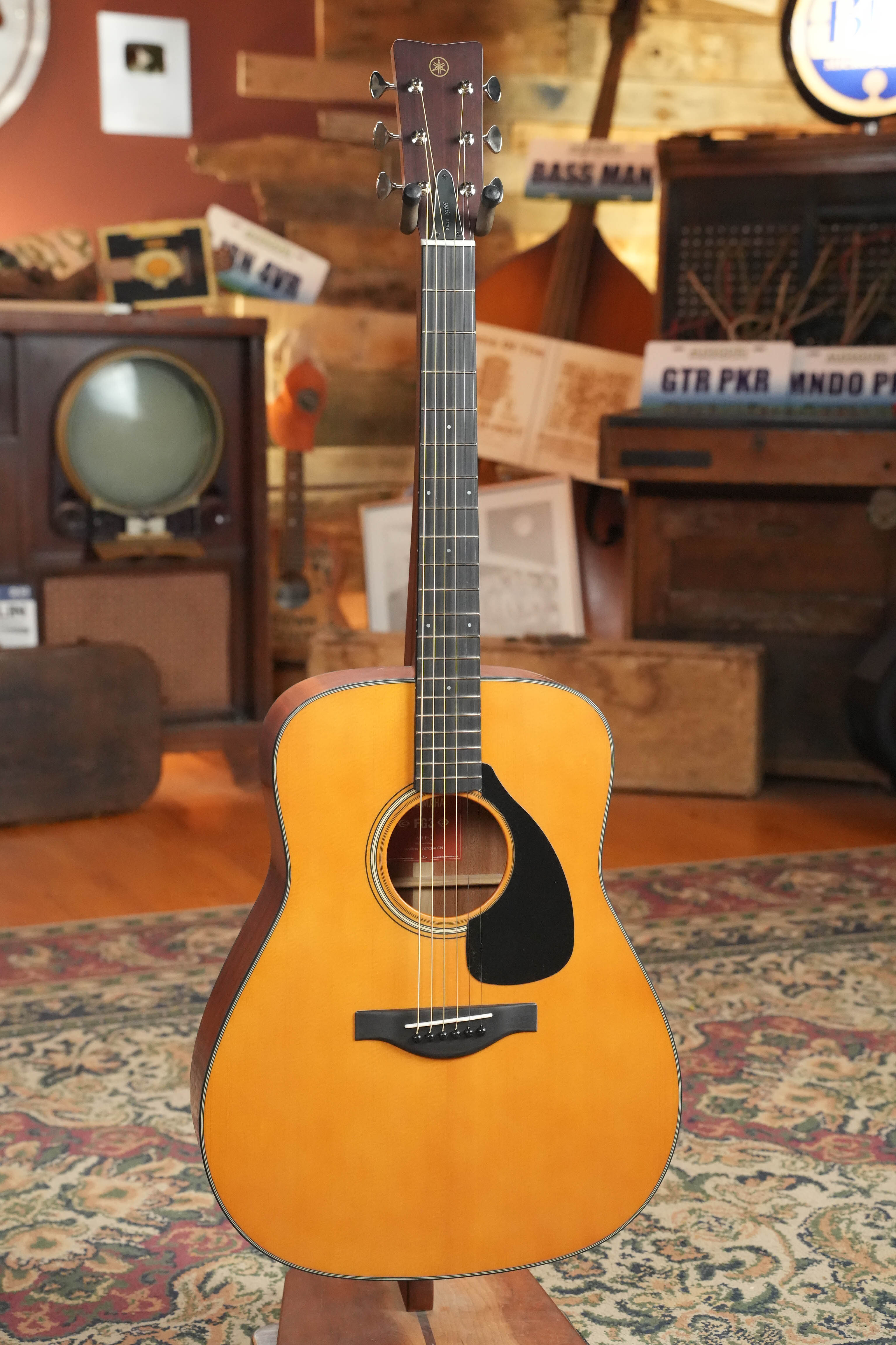 A Yamaha FG3 Red Label Dreadnought Acoustic Guitar stands upright on a patterned rug in a cozy, vintage-style room decorated with music-themed items and wood furnishings.