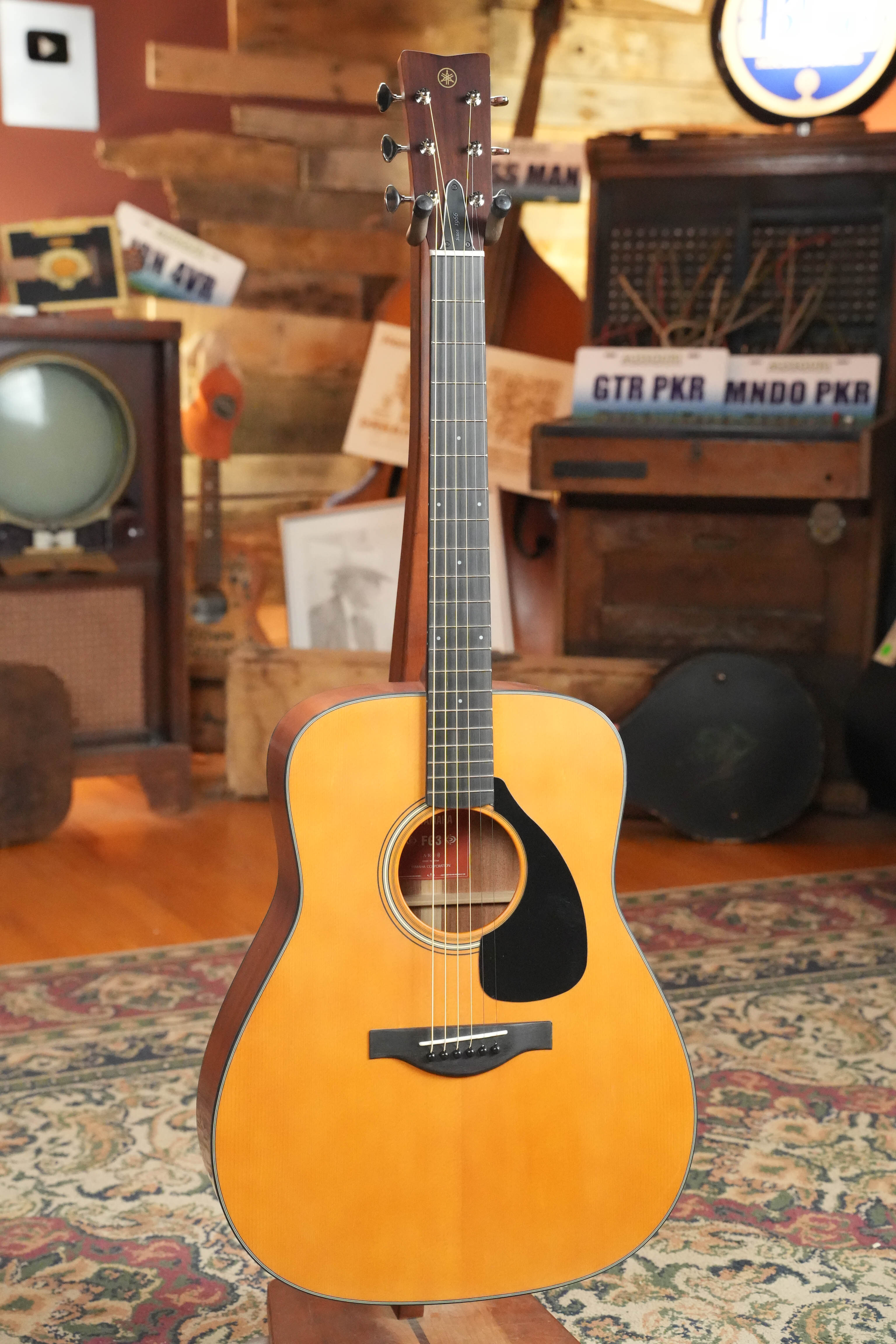 The Yamaha FG3 Red Label Dreadnought Acoustic Guitar #1210 stands upright on a patterned rug, its natural wood finish complementing vintage electronics and wood-paneled decor in the background.