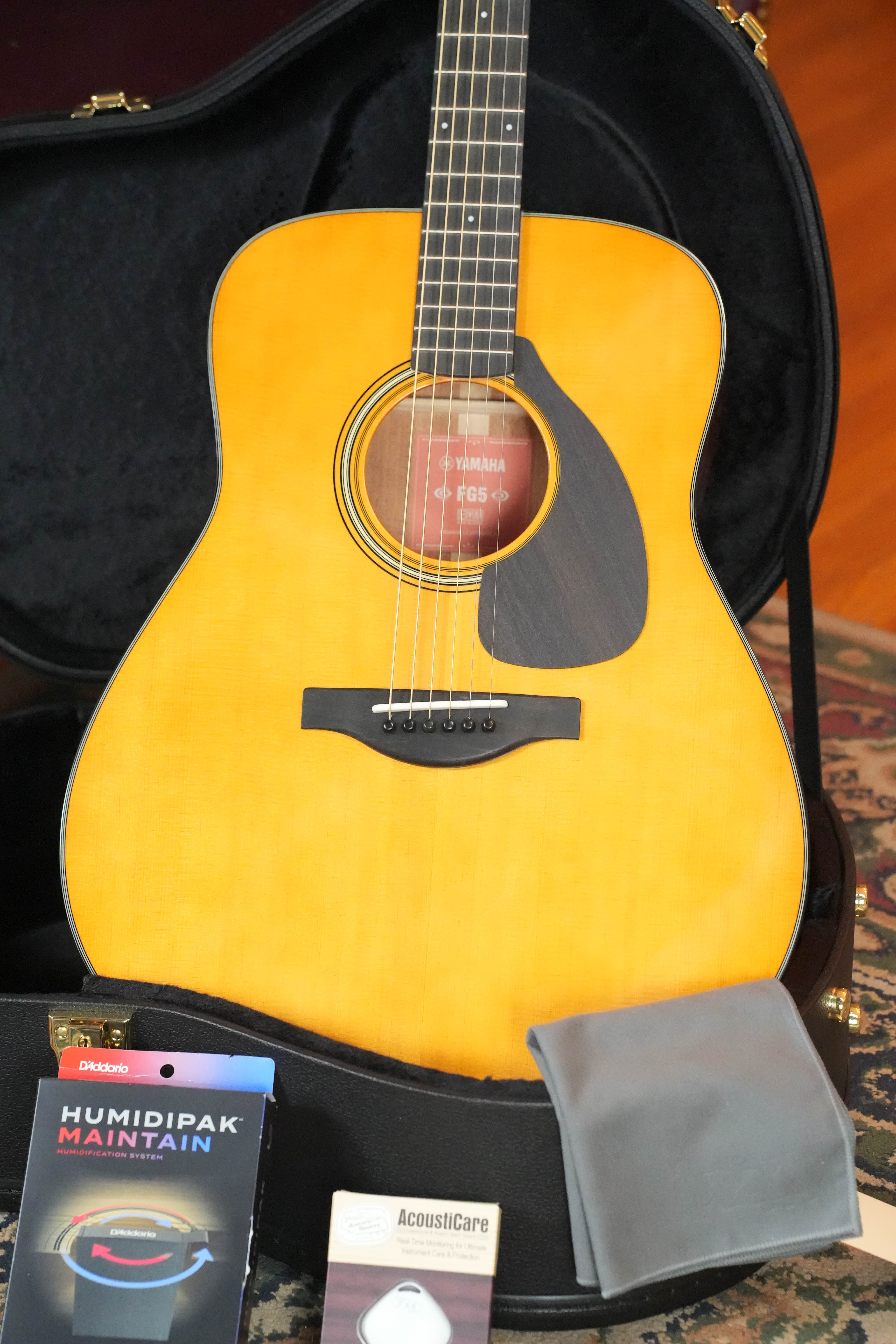 A Yamaha FG5 GL NEW Gloss Red Label Dreadnought Acoustic Guitar #227A rests in an open black case on a patterned rug, accompanied by a gray cloth, Humidipak Maintain packet, and an AcoustiCare product.