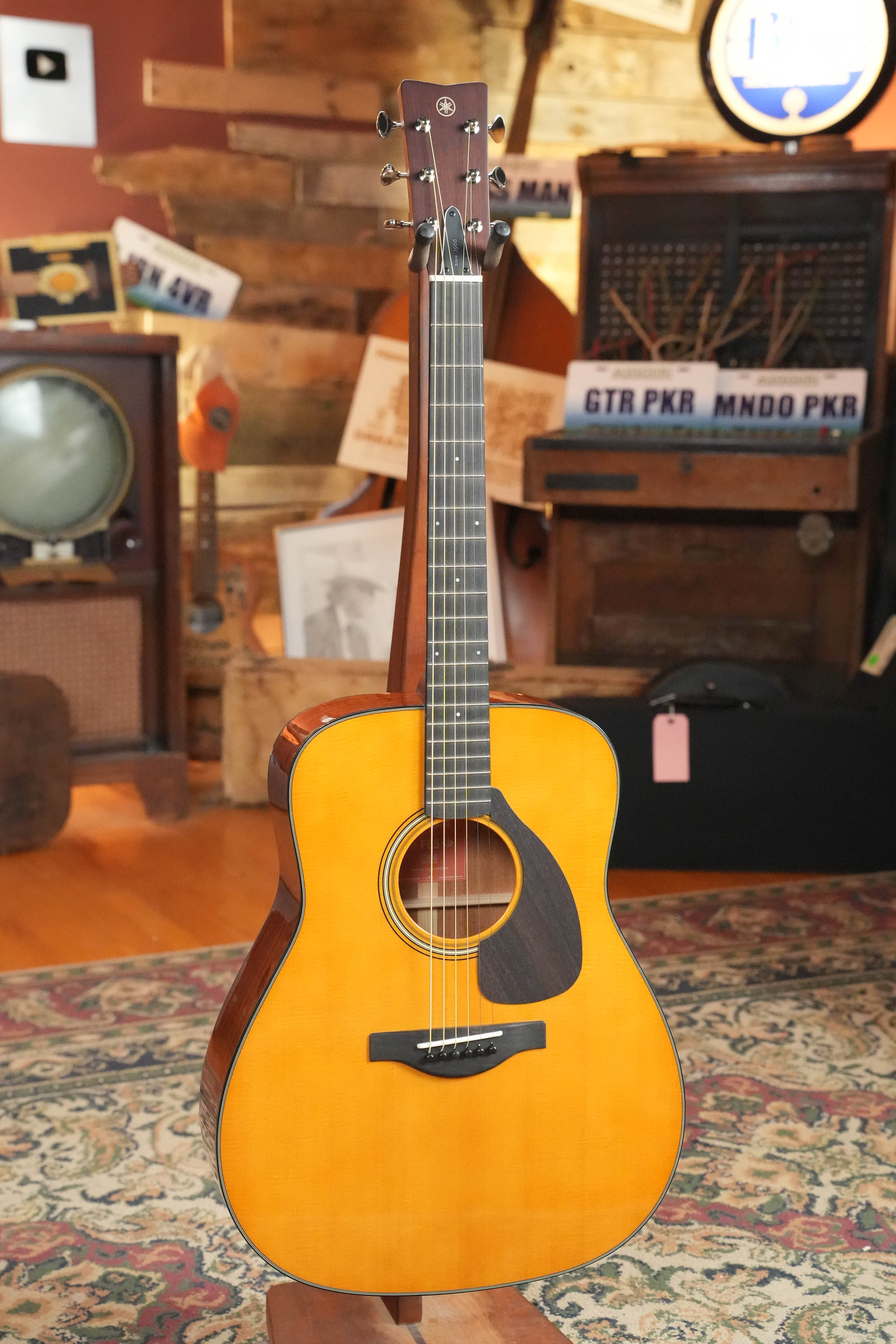 A Yamaha FG5 GL NEW Gloss Red Label Dreadnought Acoustic Guitar #227A stands on a decorative rug in a cozy room with vintage decor, including an old TV, wood-paneled walls, and framed pictures.