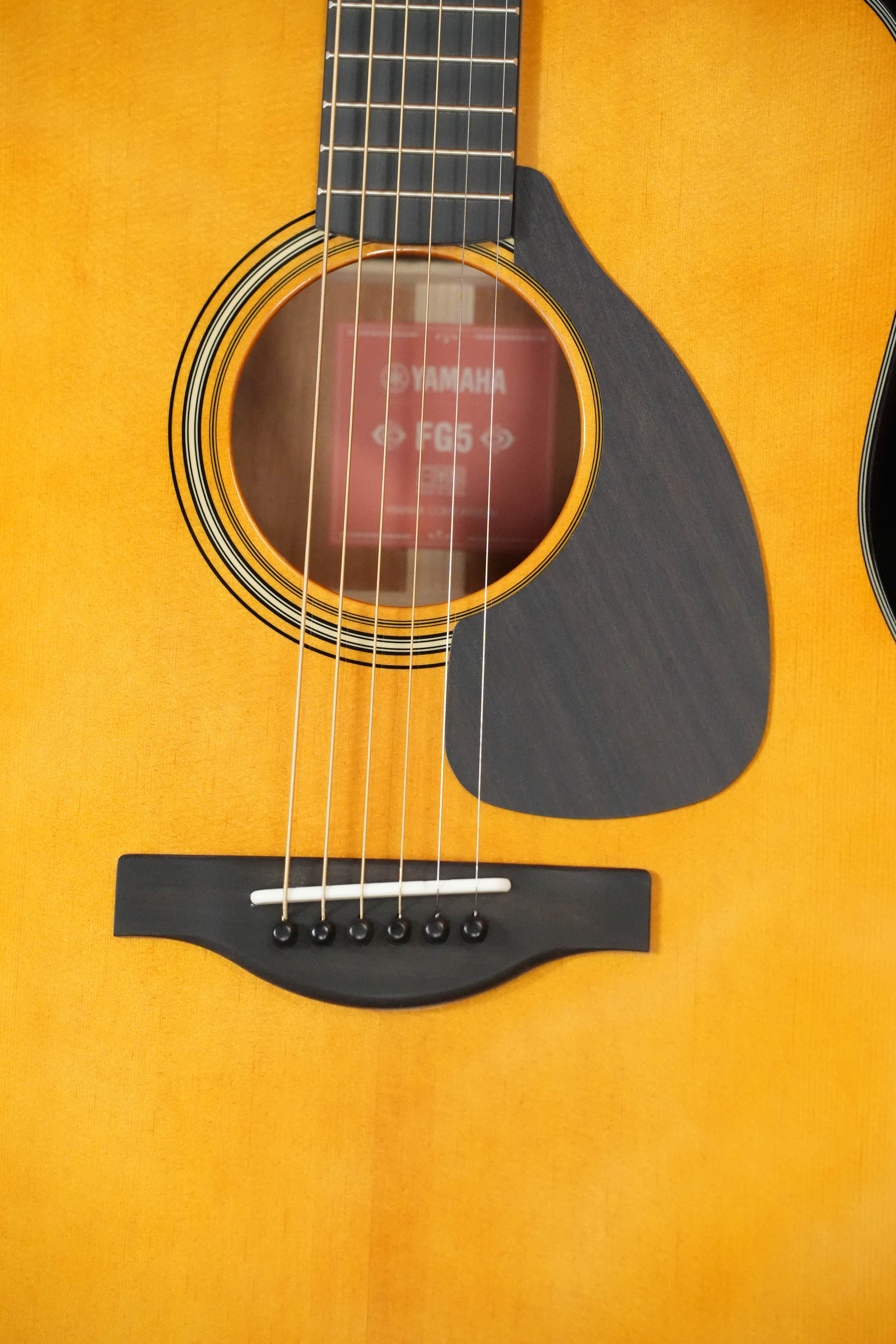 Close-up of the Yamaha FG5 GL NEW Gloss Red Label Dreadnought Acoustic Guitar #732A, showing its solid spruce top, visible strings, sound hole, black pickguard, and label reading Yamaha FG5. Features Acoustic Resonance Enhancement. Brand: Yamaha.