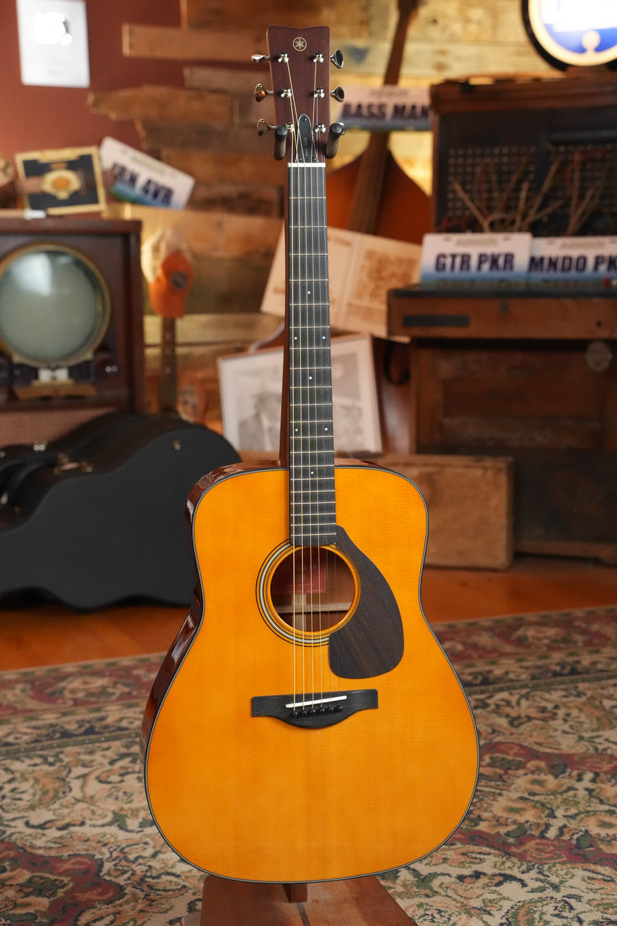 A Yamaha FG5 GL NEW Gloss Spruce/Mahogany Red Label Dreadnought Acoustic Guitar #321A stands on a patterned rug amid vintage gear in a cozy, rustic room, its Acoustic Resonance Enhancement enriching each note.