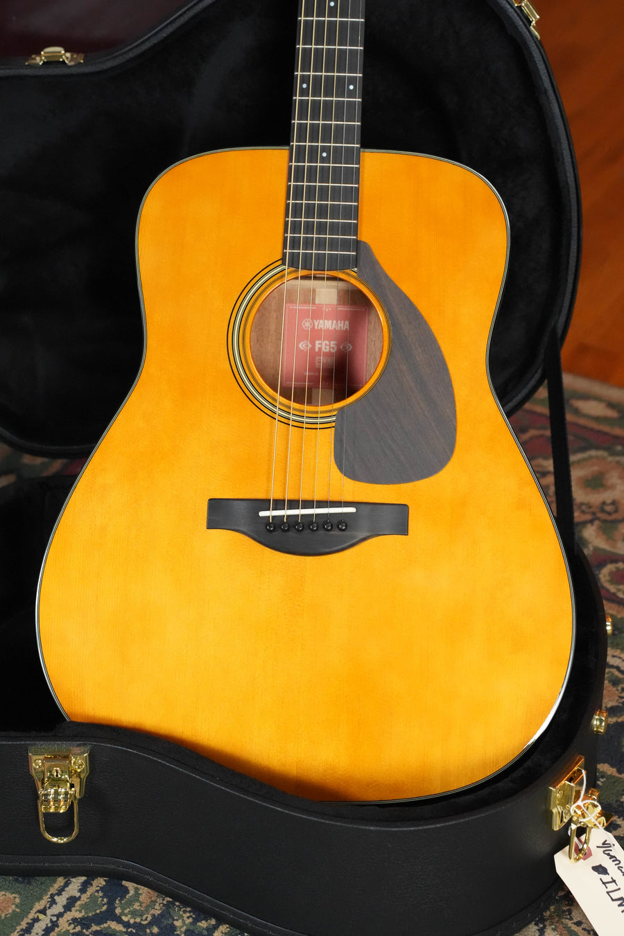 A Yamaha FG5 GL NEW Gloss Spruce/Mahogany Red Label Dreadnought Acoustic Guitar (#520A) with a solid spruce top and glossy finish rests in an open black hard case on a patterned rug, featuring a dark pickguard.