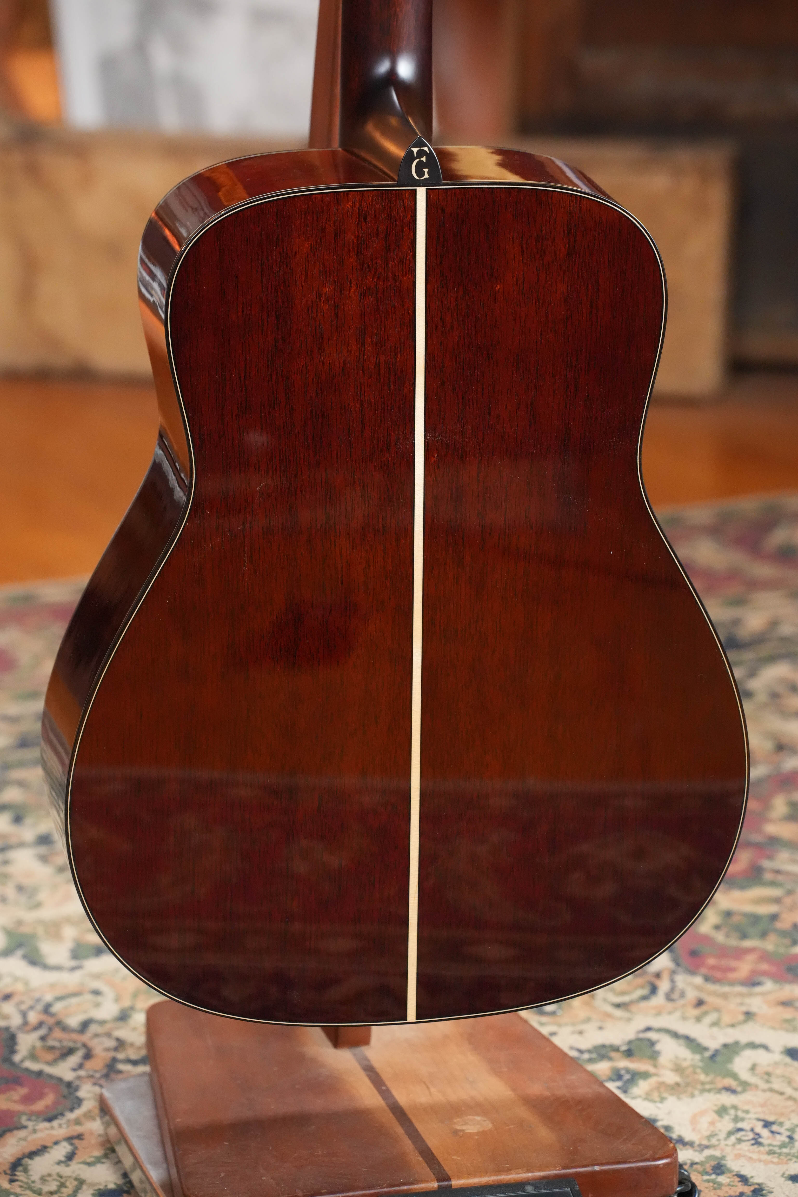 A close-up of the back of a Yamaha FG9 M Adirondack/Mahogany Dreadnought Acoustic Guitar #112A on a wooden stand over a patterned rug, highlighting its glossy wood finish with a center stripe and renowned rich acoustic tone.