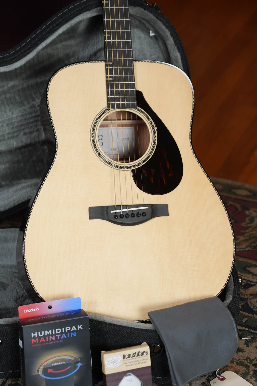 The Yamaha FG9 M Adirondack/Mahogany Dreadnought Acoustic Guitar #112A is shown in a hard case with accessories, including a Humidipak, AcousticCare product, and gray cloth, showcasing its premium build and rich acoustic tone.