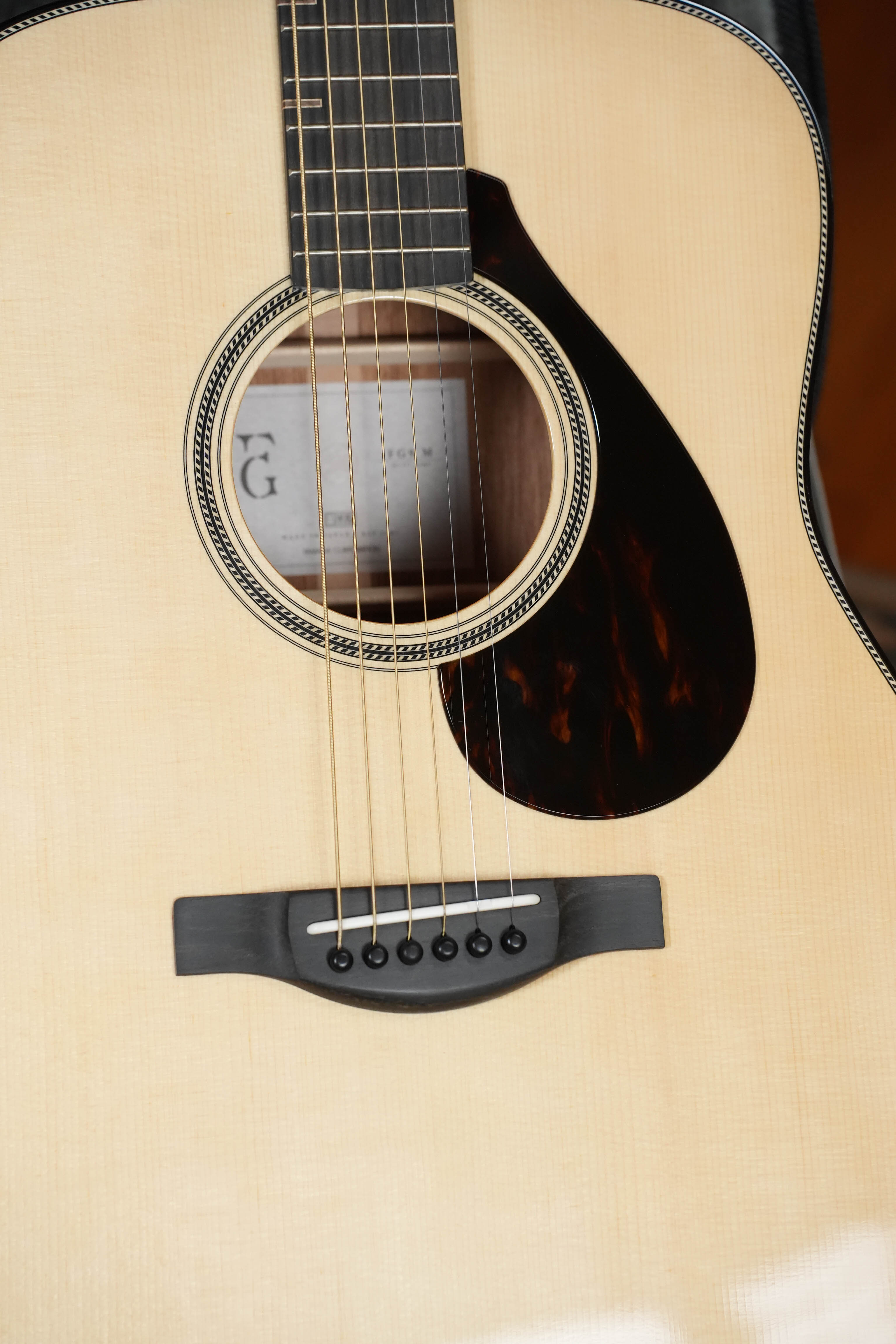 Close-up of the Yamaha FG9 M Adirondack/Mahogany Dreadnought Acoustic Guitar #112A, showing its strings, wooden bridge, pickguard, and light wood finish. The Adirondack spruce top and interior label are visible through the sound hole.
