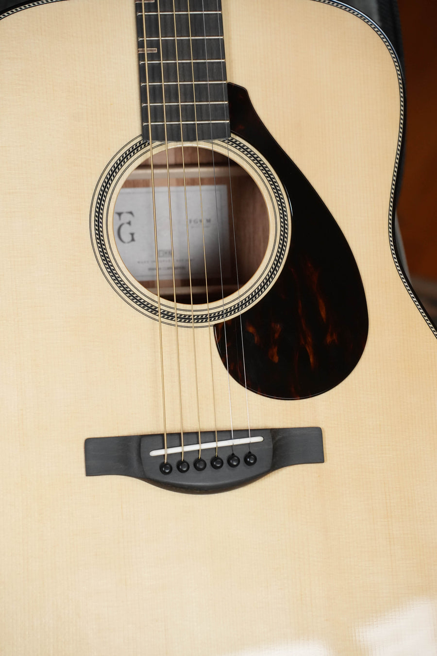 Close-up of the Yamaha FG9 M Adirondack/Mahogany Dreadnought Acoustic Guitar #112A, showing its strings, wooden bridge, pickguard, and light wood finish. The Adirondack spruce top and interior label are visible through the sound hole.