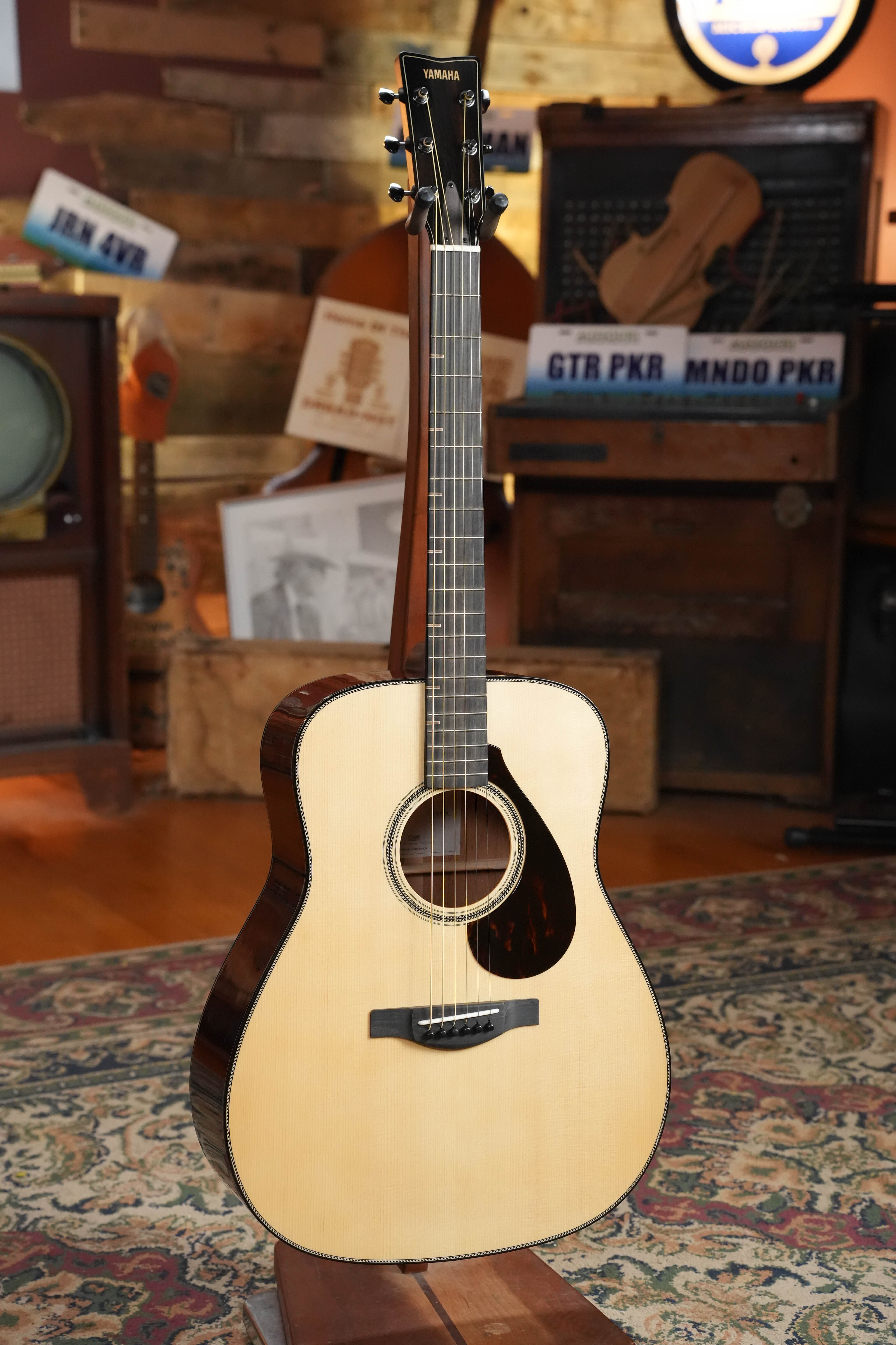 A Yamaha FG9 M Adirondack/Mahogany Dreadnought Acoustic Guitar #112A stands upright on a stand in a cozy, vintage-style room with musical decor and a patterned rug.