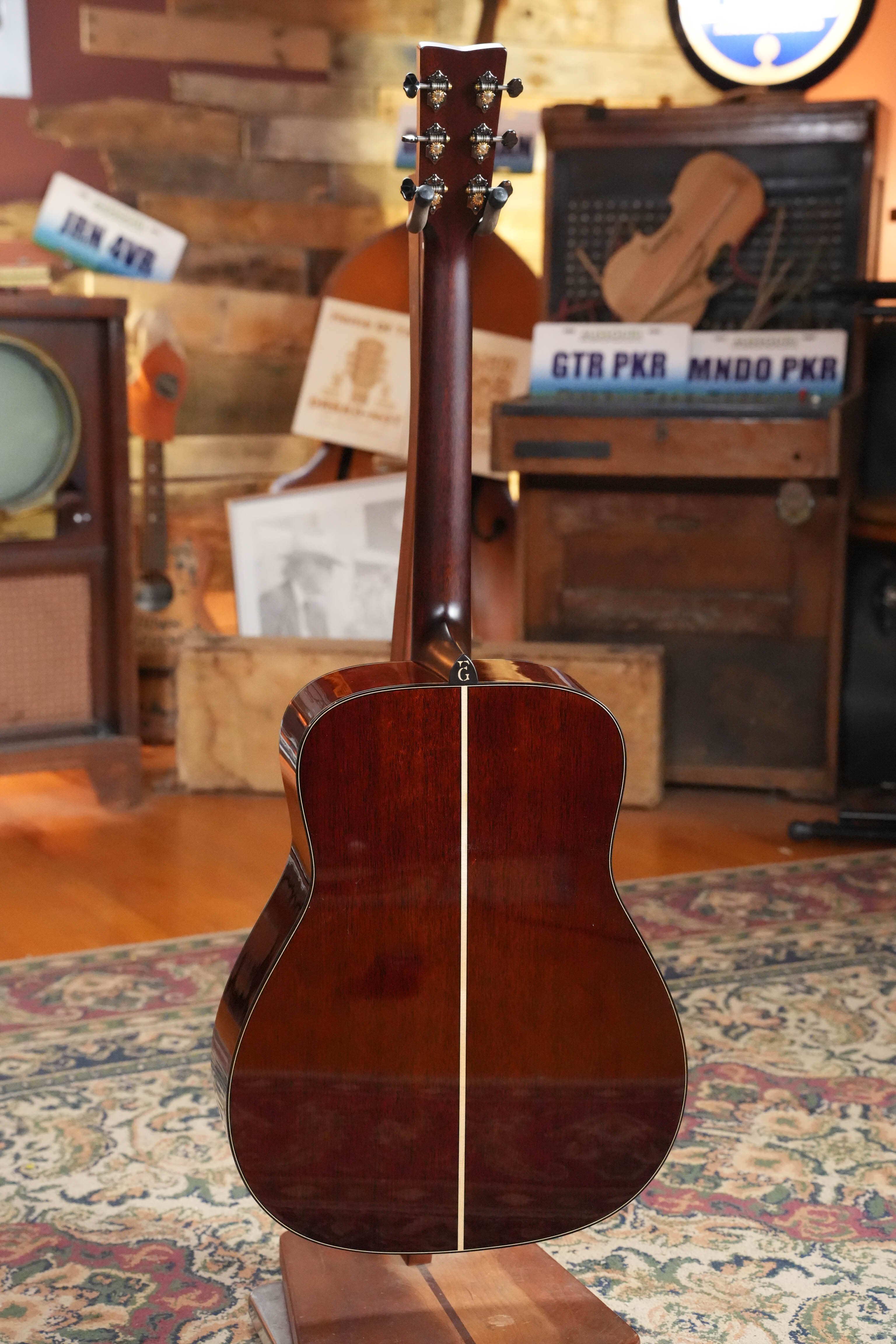 The Yamaha FG9 M Adirondack/Mahogany Dreadnought Acoustic Guitar #112A by Yamaha is displayed from the back on a wooden stand in a cozy, vintage-inspired room. Warm lighting and a patterned carpet highlight its inviting acoustic appeal.